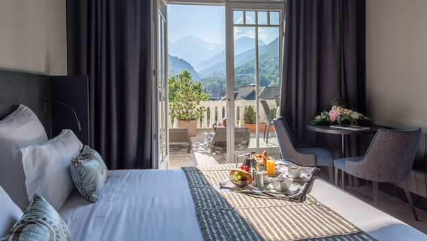 A hotel room with a bed, a breakfast tray, a table with chairs, and a view of mountains through a large window. - Golf Hôtel (Brides-les-Bains)