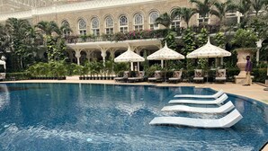 Une piscine entourée de chaises longues et de parasols, elle-même bordée d’une végétation luxuriante et d’une façade de style classique.