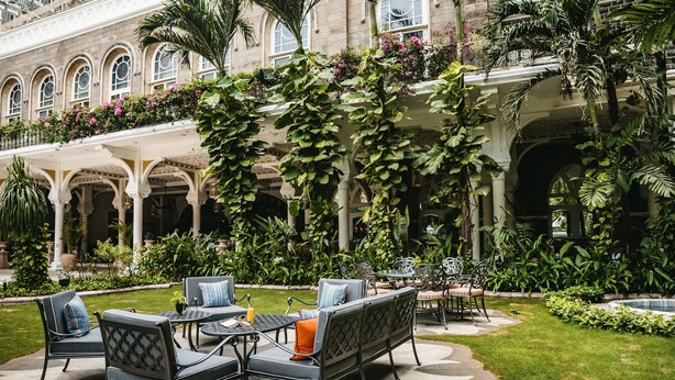 Une terrasse extérieure aménagée avec des sièges et des tables, entourée d’une végétation luxuriante et bordée d’un bâtiment historique doté d’arcades et de colonnes.