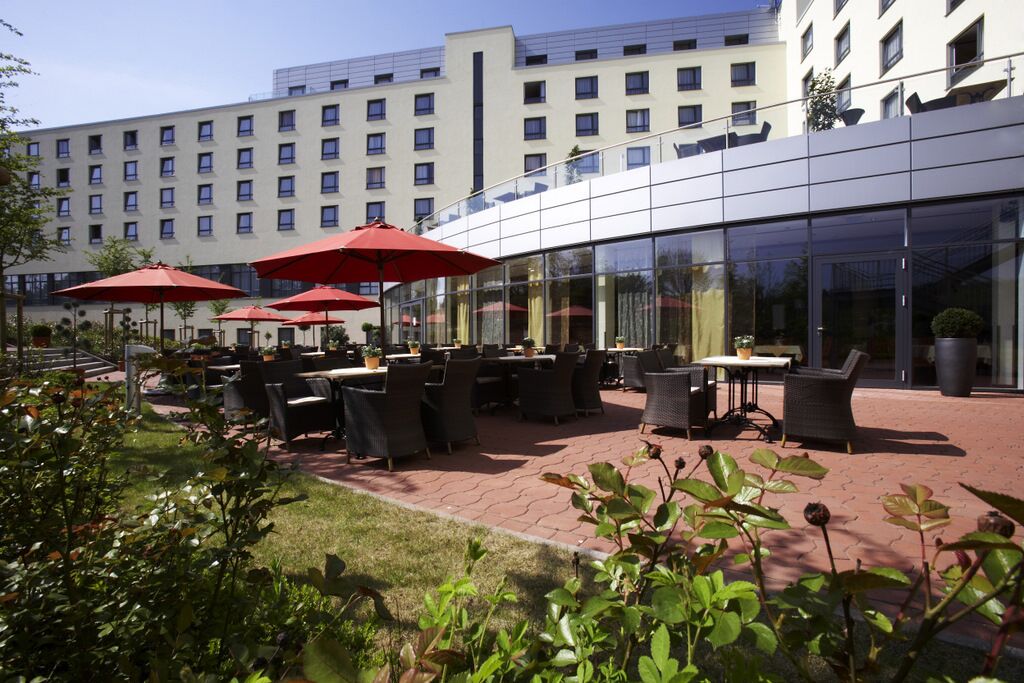 An outdoor patio area with tables and chairs, red umbrellas, and a modern building in the background.