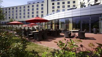 An outdoor patio area with tables and chairs, red umbrellas, and a modern building in the background.