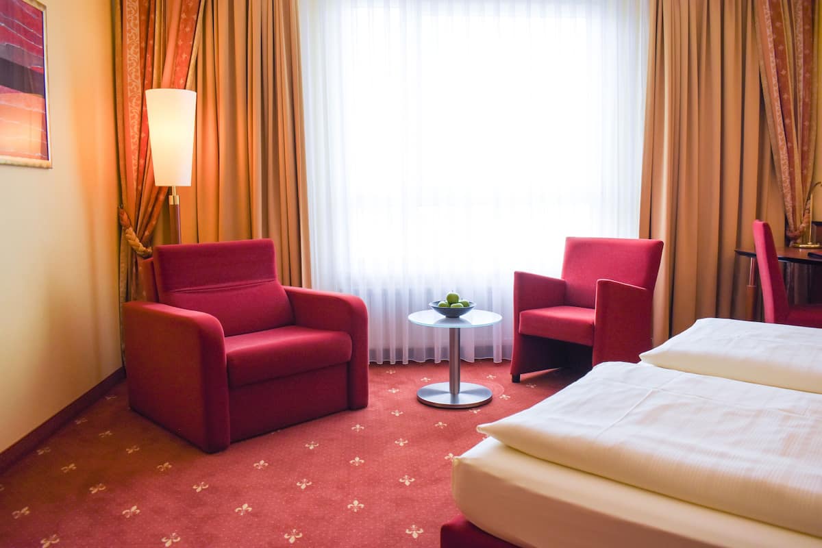 A hotel room with a bed, two red armchairs, a small round table with a bowl of fruit, and a lamp.