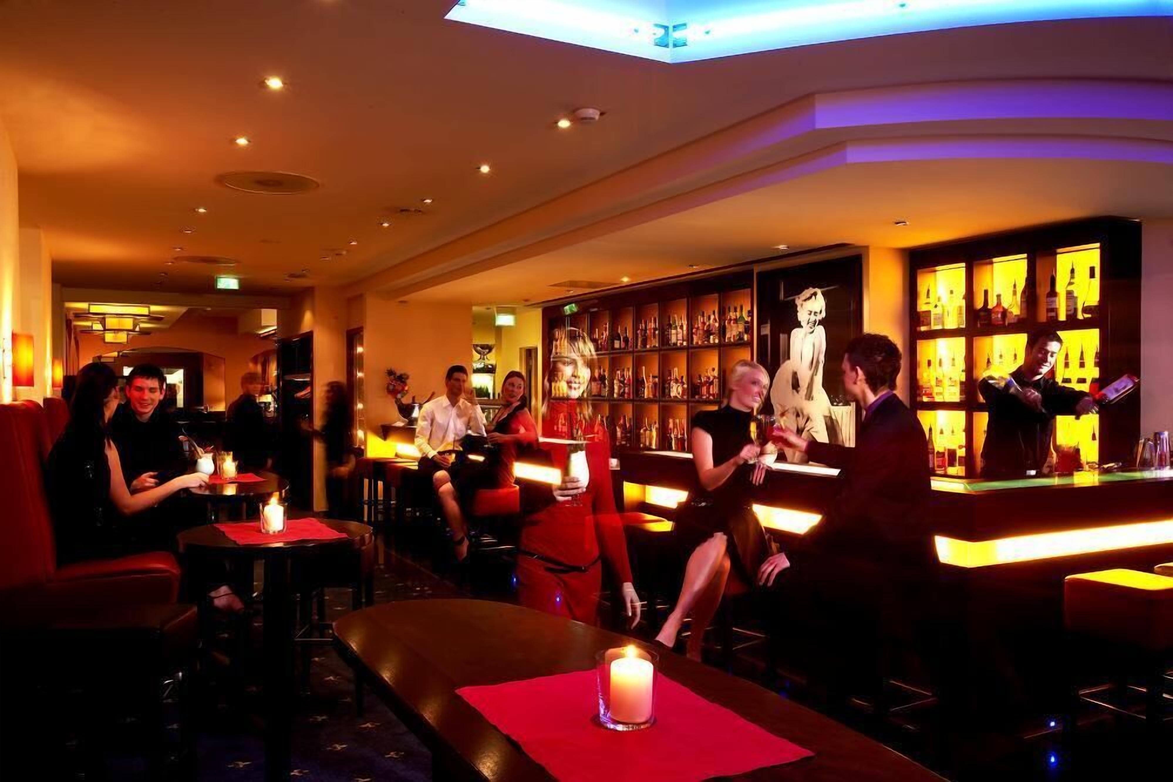 Eine Bar mit Tischen, Stühlen und einer Theke. Personen sitzen und stehen dort, und ein Barkeeper ist anwesend.