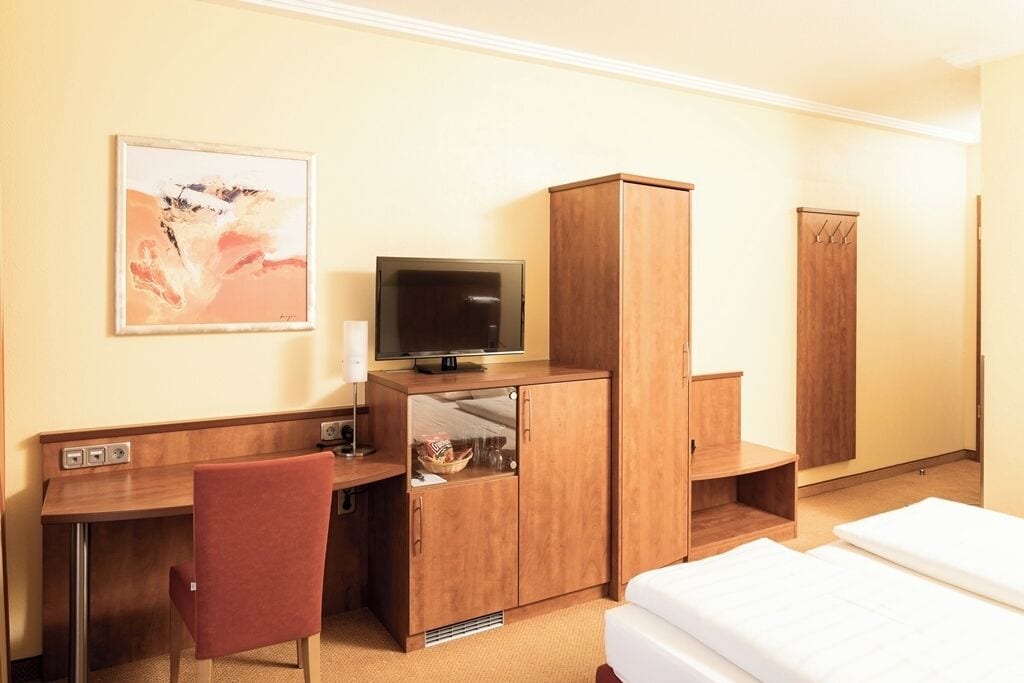 A hotel room with a wooden desk, a television, a wardrobe, a bed, and a painting on the wall.