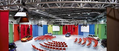 A spacious indoor exhibition hall with a high ceiling and metal beams, featuring rows of red chairs and several cars on display.