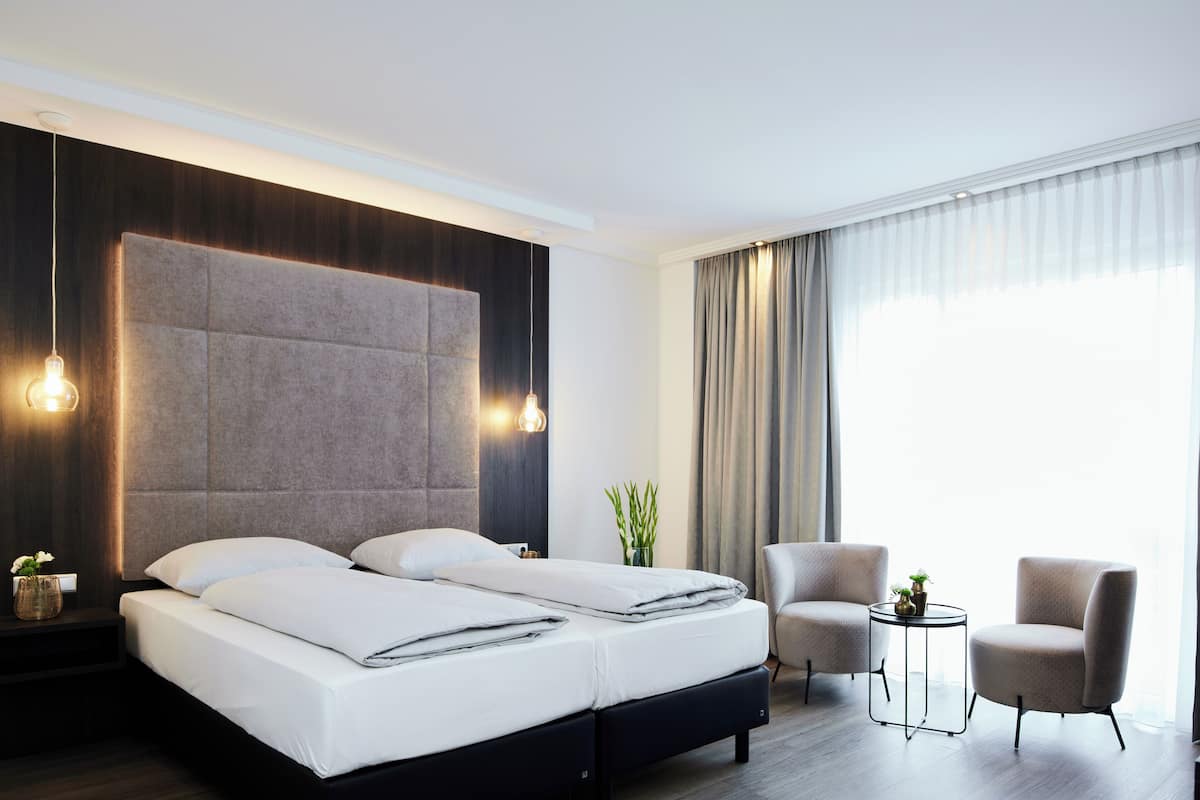 Une chambre d’hôtel moderne, dotée d’un grand lit, de deux fauteuils, d’une petite table d’appoint et d’une grande fenêtre avec des rideaux.