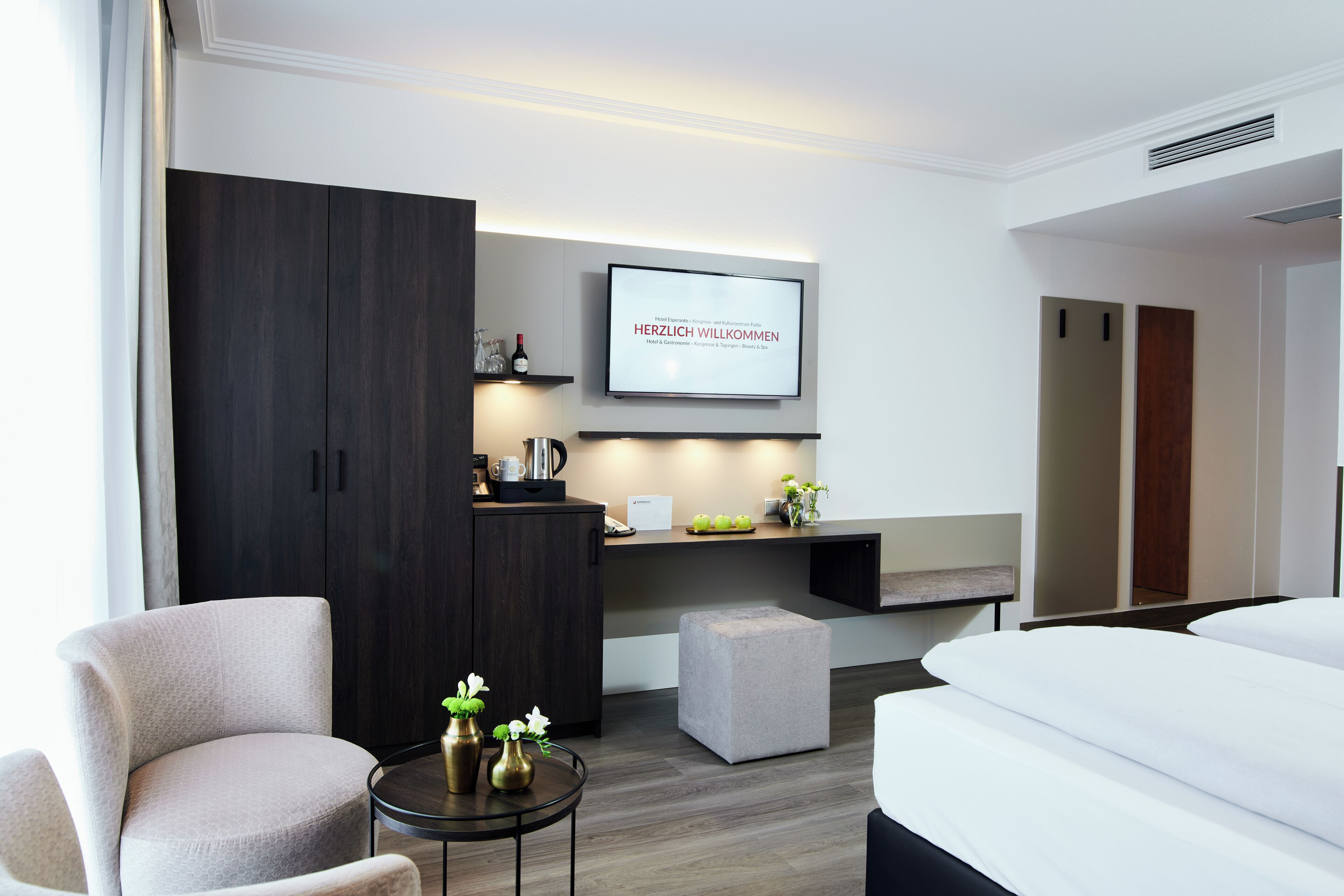 A modern hotel room with a bed, a desk, a chair, a small table with decorative items, and a television displaying a welcome message.