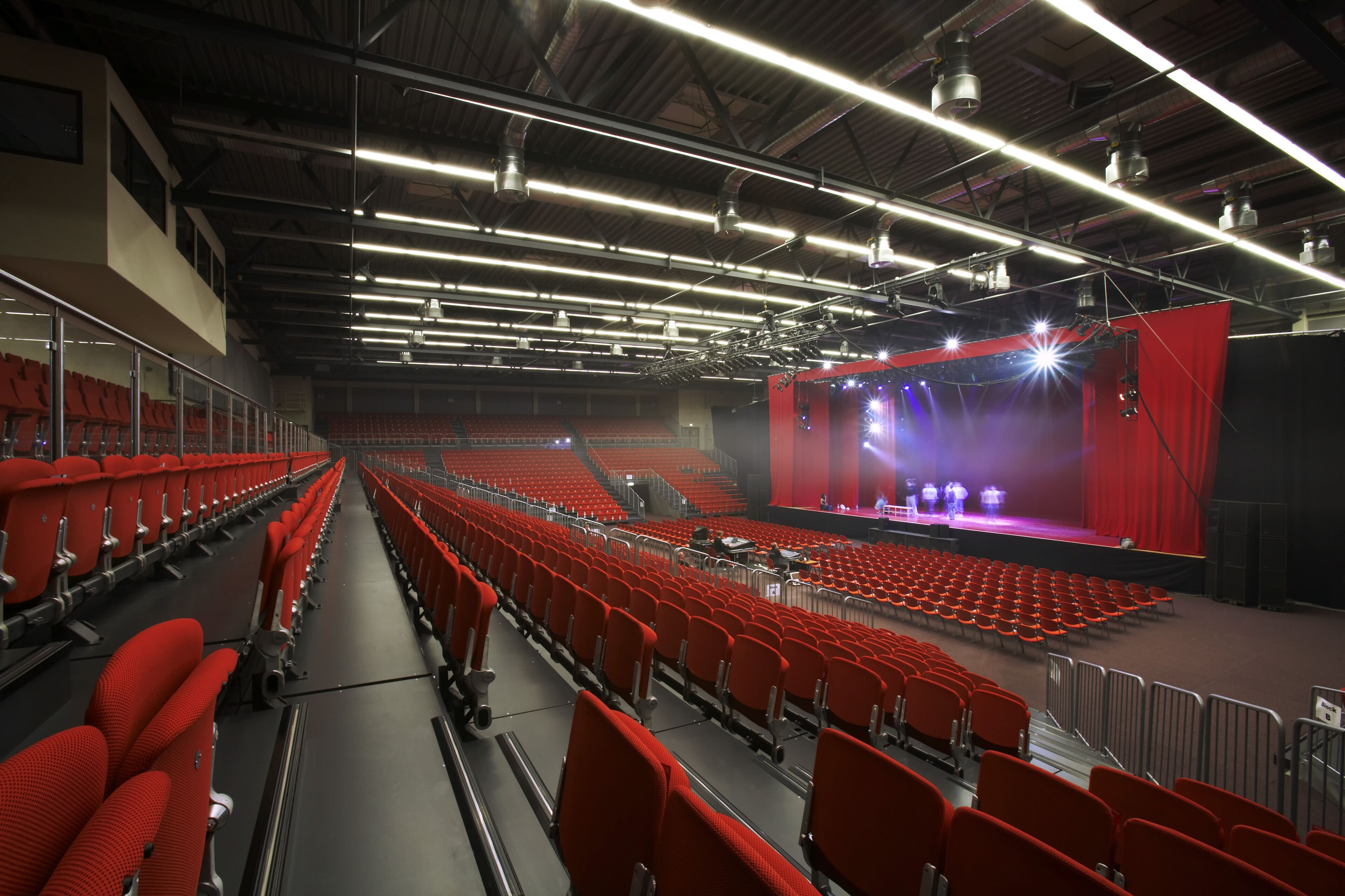 An indoor auditorium with rows of red seats, a stage with a red curtain, and overhead lighting.