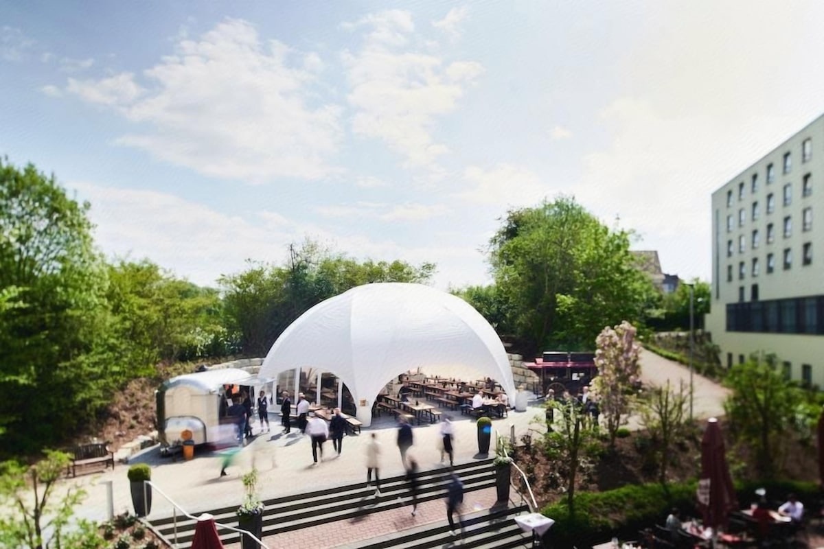 An outdoor dining area with a large white dome structure, surrounded by greenery and a building in the background.