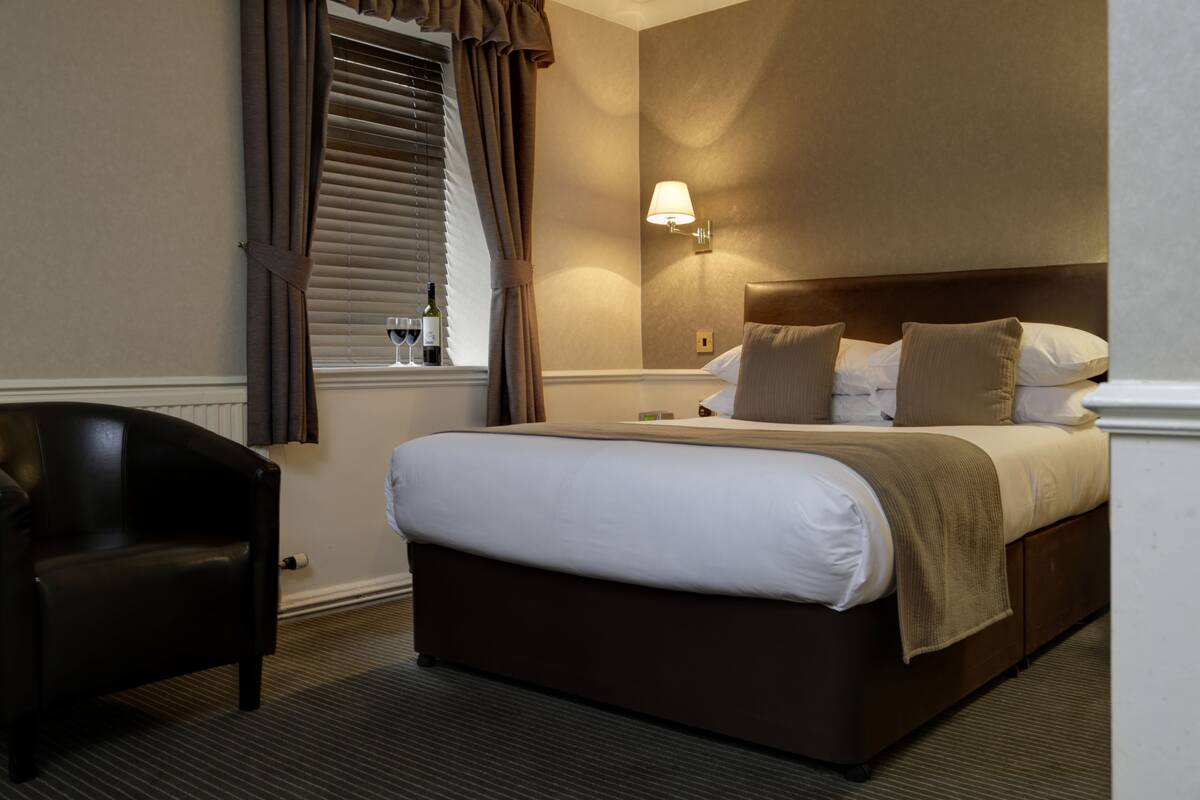 A hotel room with a large bed, a nightstand, a lamp, a chair, and a window with curtains.