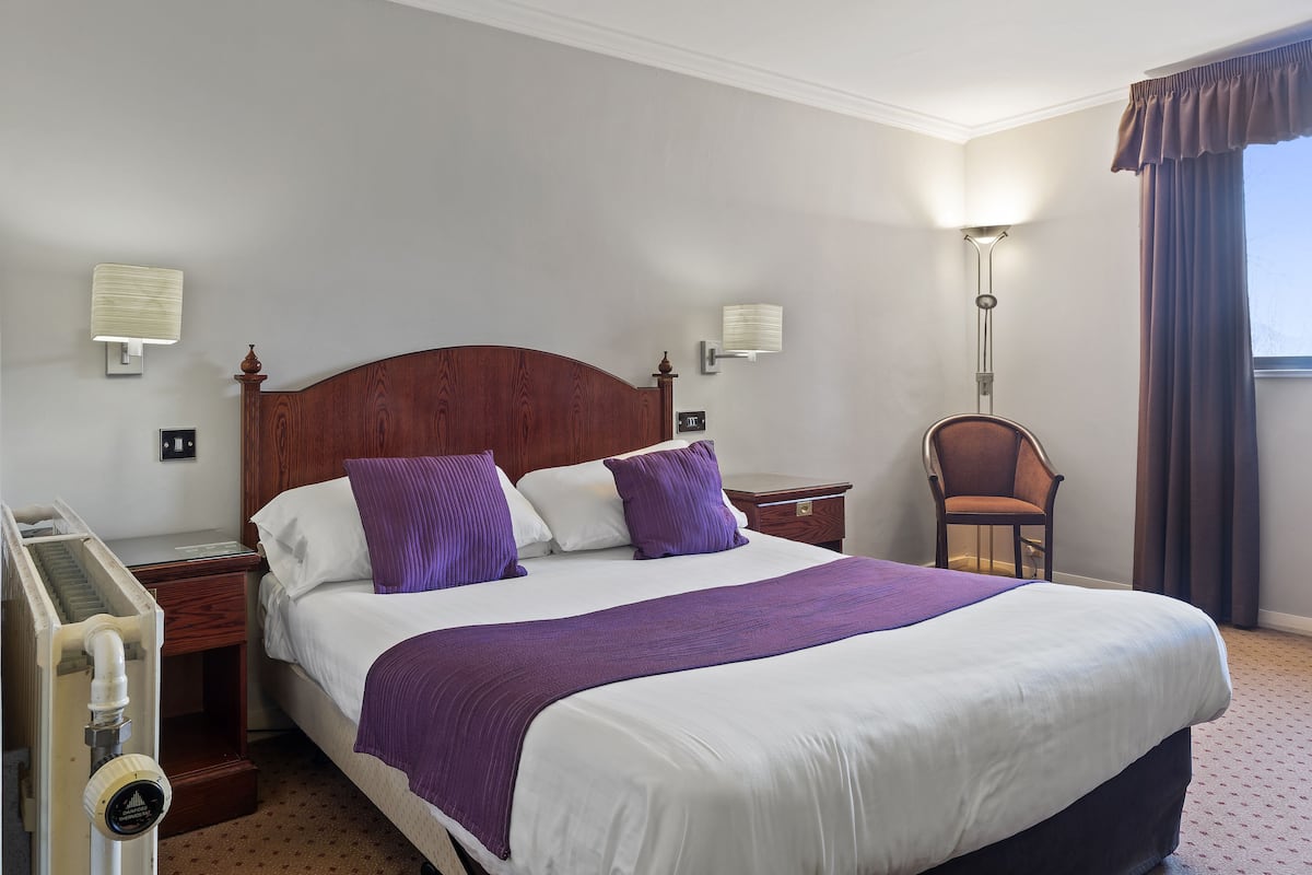A hotel room with a large bed, two bedside tables, a chair, a lamp, and a window with curtains.