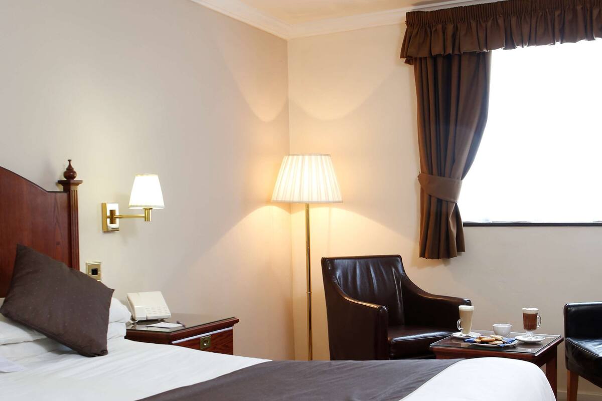 A hotel room with a large bed, a desk, a chair, a lamp, and a window with curtains.