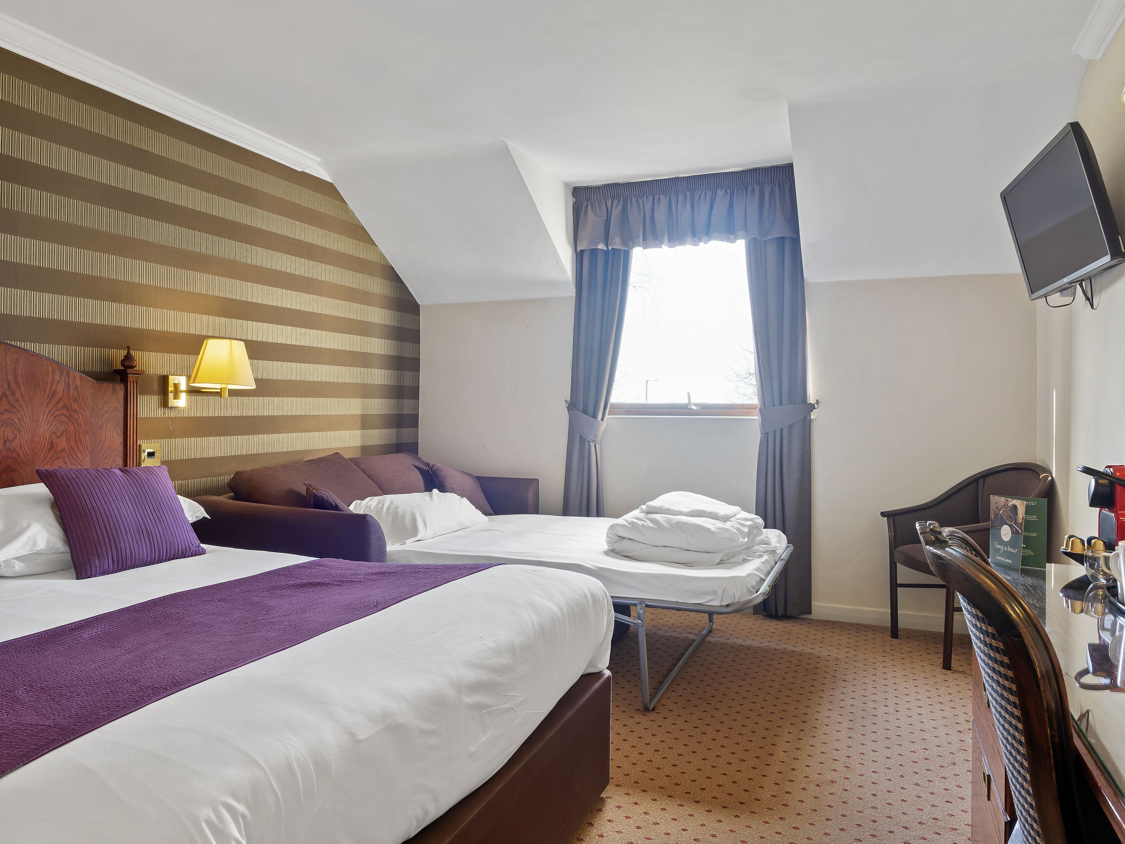 A hotel room with a large bed, a smaller bed, a desk with a computer, a lamp, and a window with curtains.