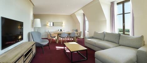 A modern hotel room with a flat-screen TV, a sofa, a dining table with chairs, and a large window with curtains.