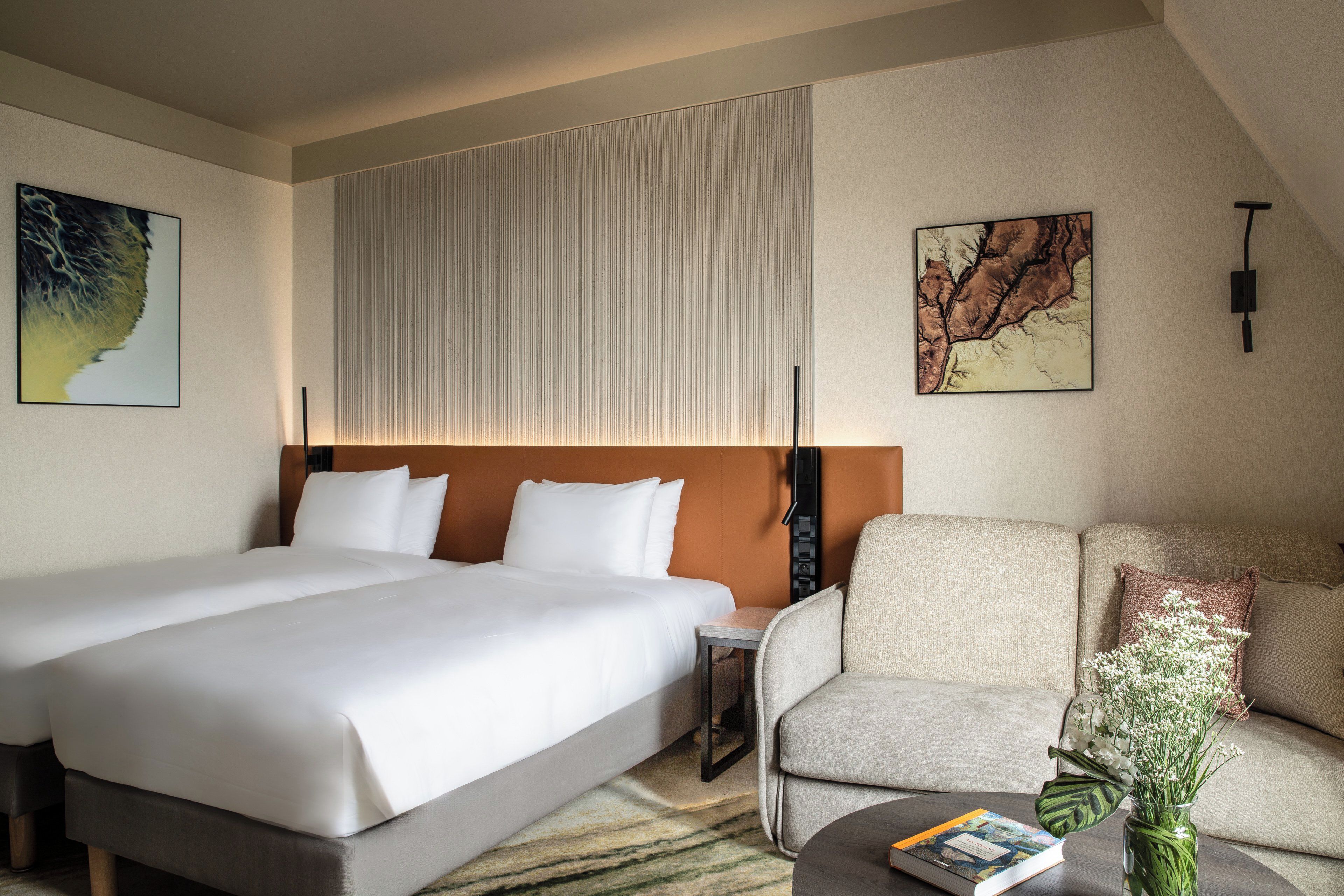 Une chambre d’hôtel avec un grand lit, un canapé, une table basse avec un livre et des fleurs, et deux œuvres d’art accrochées au mur.