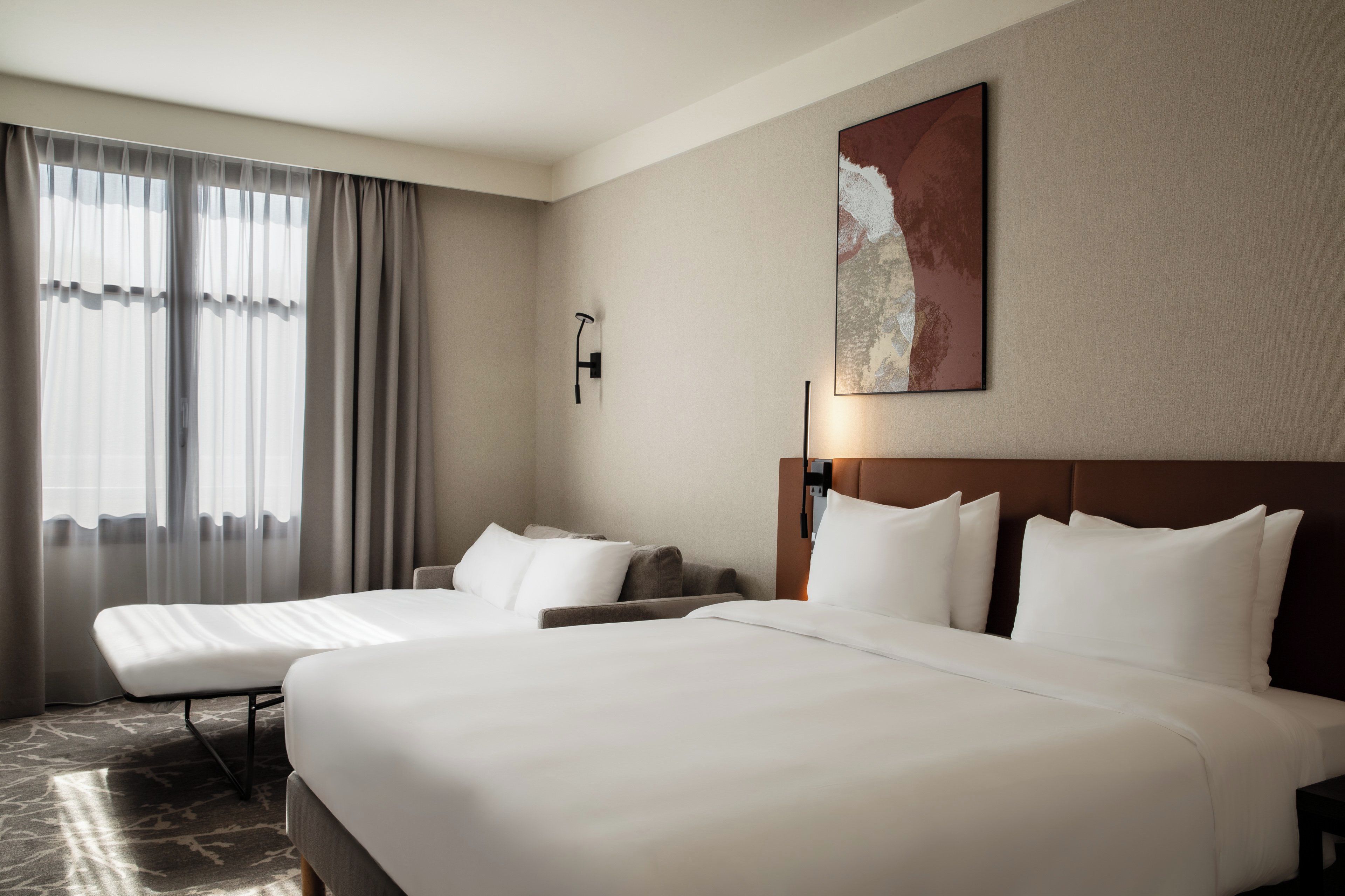 Quarto de hotel com duas camas, uma janela grande com cortinas, uma luminária na parede e um quadro.