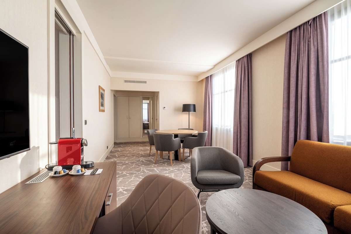 Een hotelkamer met een televisie, een koffieapparaat, een eettafel met stoelen, een bank en een groot raam met gordijnen.