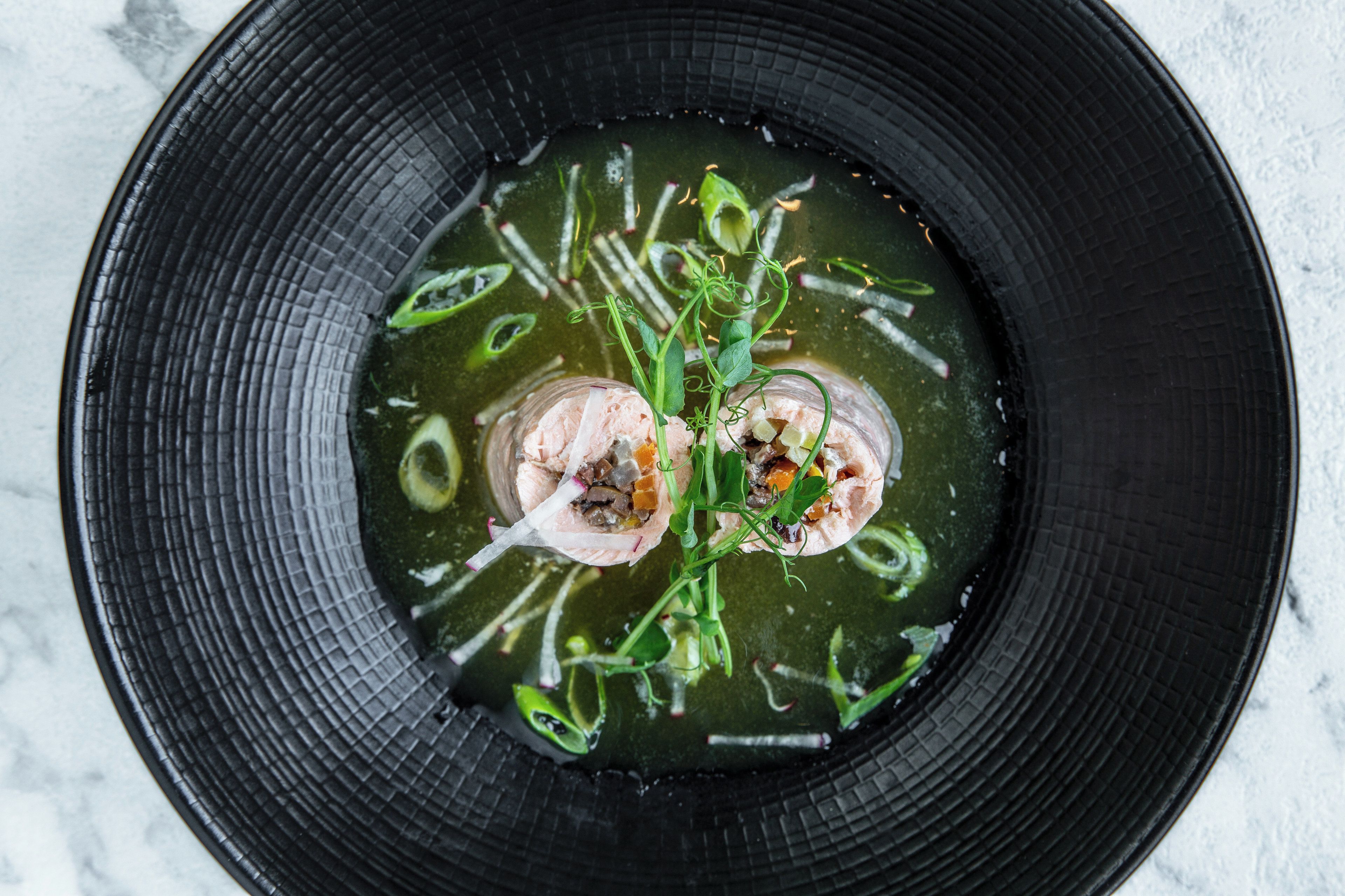 Ein schwarzer Teller mit Suppe, grünem Gemüse und einem Stück Fleisch.