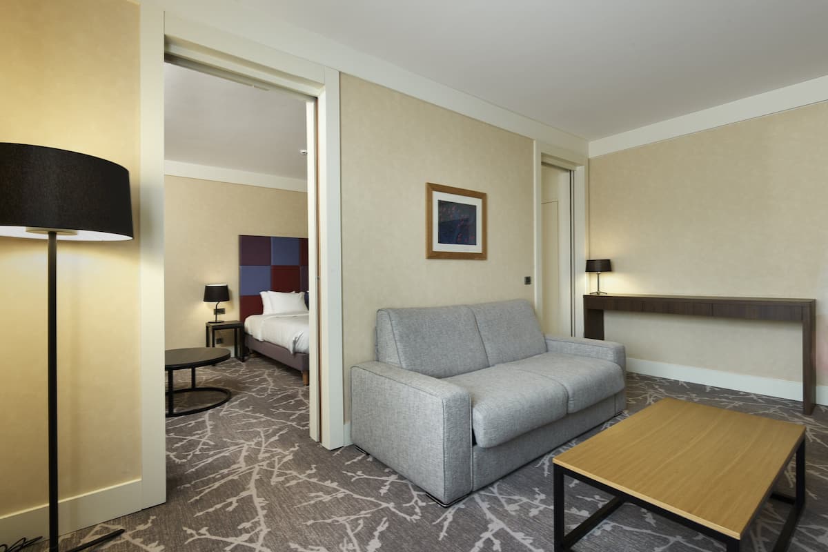 A hotel room with a grey sofa, a wooden coffee table, a small round table with a lamp, and a bed with a headboard.