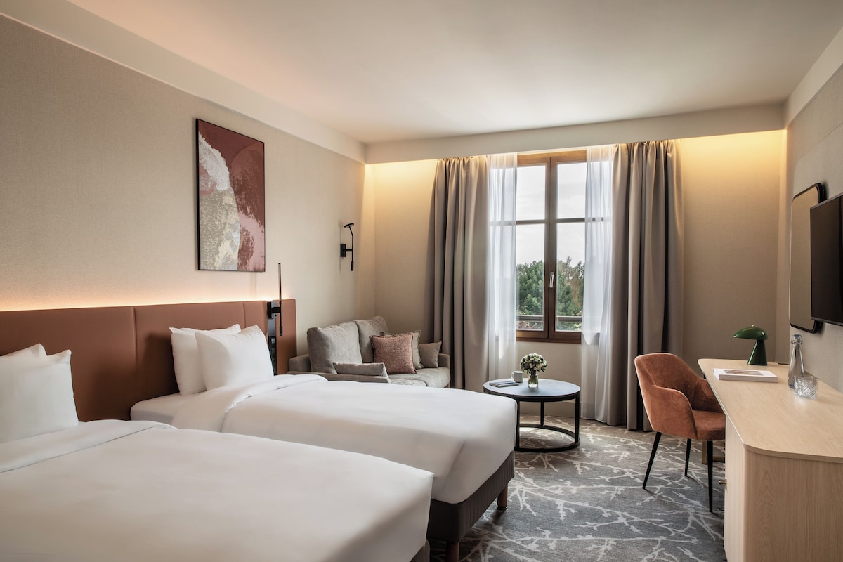 A hotel room with two beds, a desk, a chair, a small table, and a window with curtains.