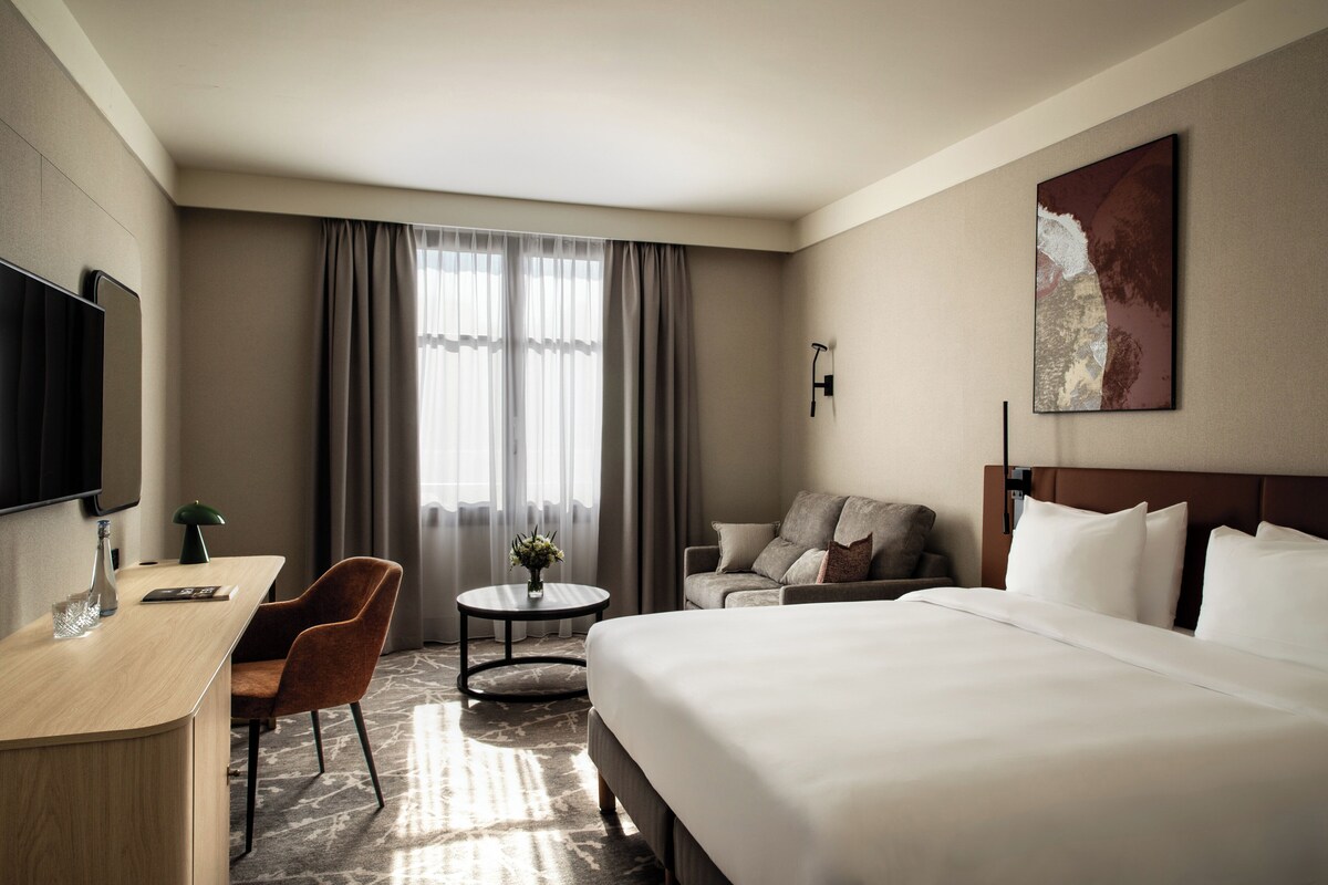 Una camera d'albergo con un letto, una scrivania, una sedia, un tavolino con un vaso e un'ampia finestra con tende.