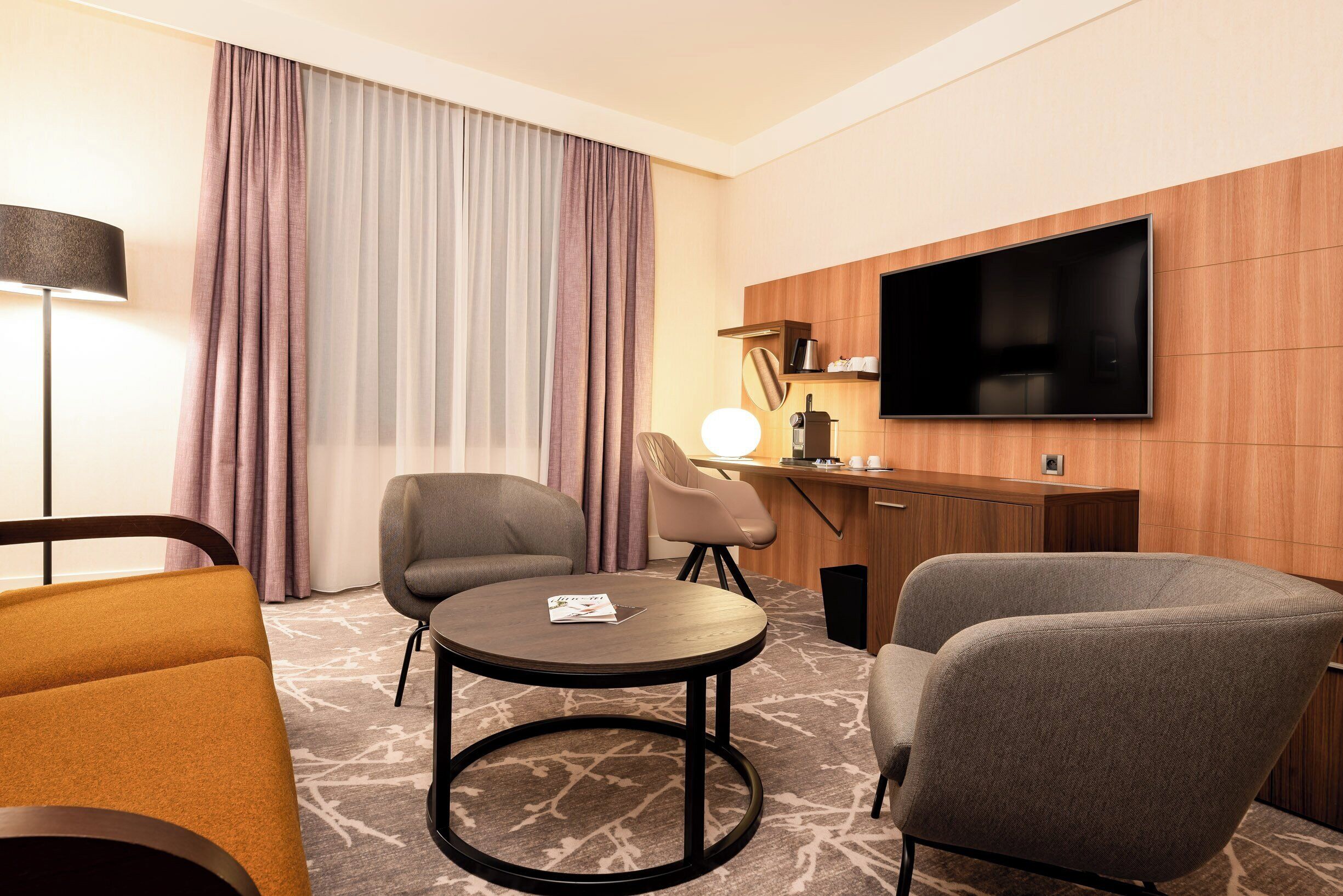 A hotel room with a TV, two armchairs, a small round table, and a desk with a lamp.