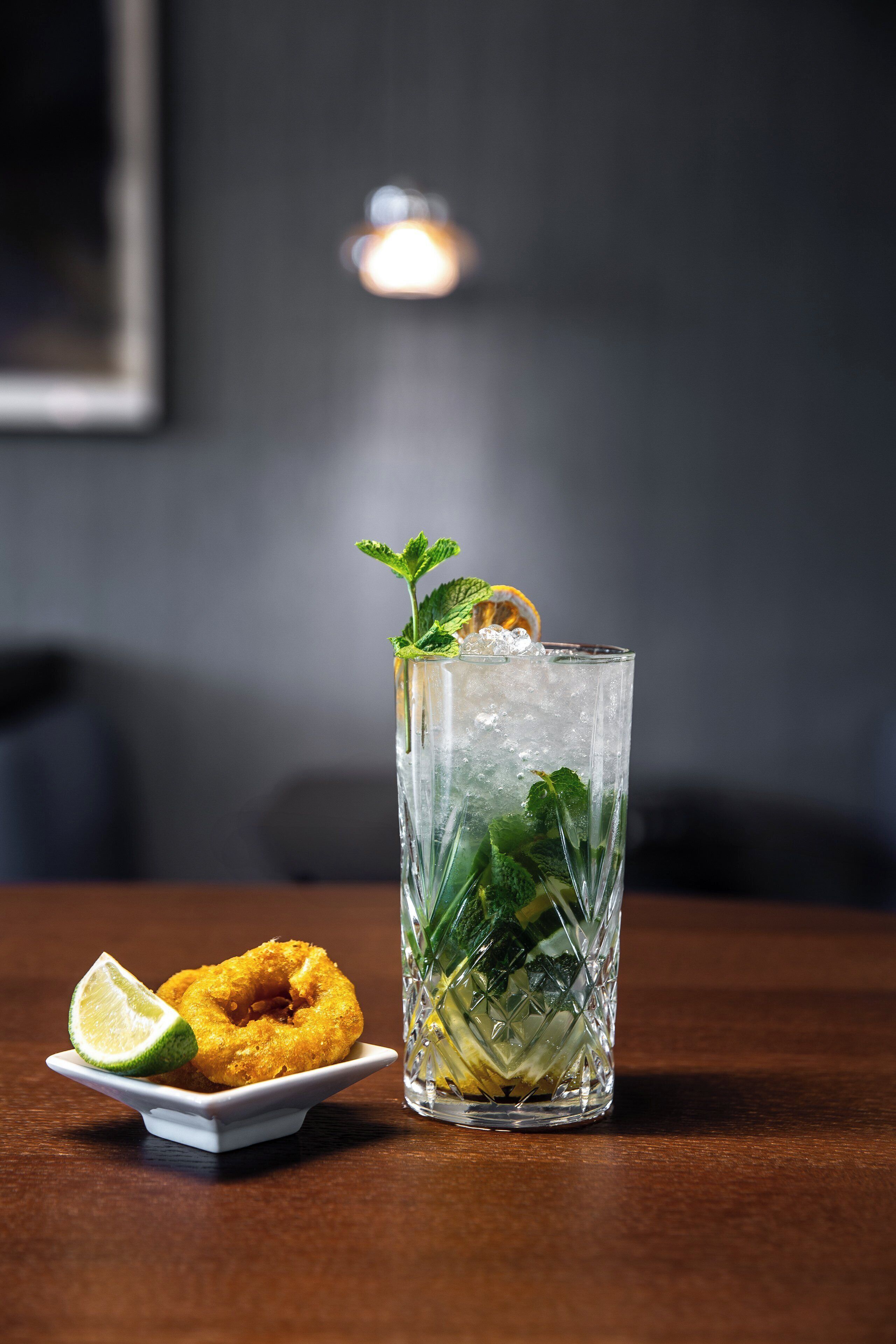 Un bicchiere di bevanda ghiacciata con menta e limone, accompagnato da una pasta fritta e una fetta di lime su un tavolo di legno.