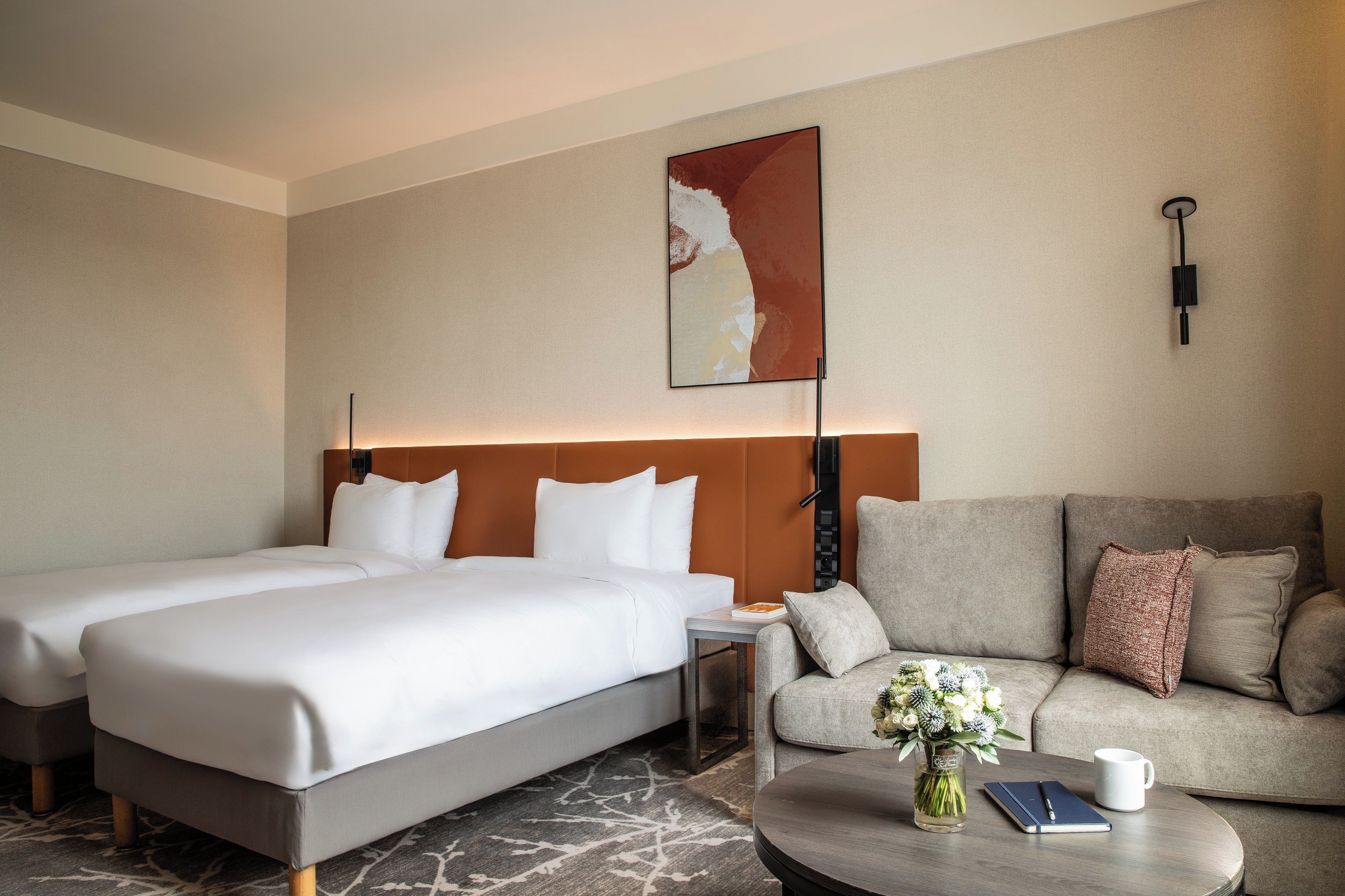 A hotel room with a large bed, a sofa, a small table with a vase and a notebook, and a wall-mounted lamp.