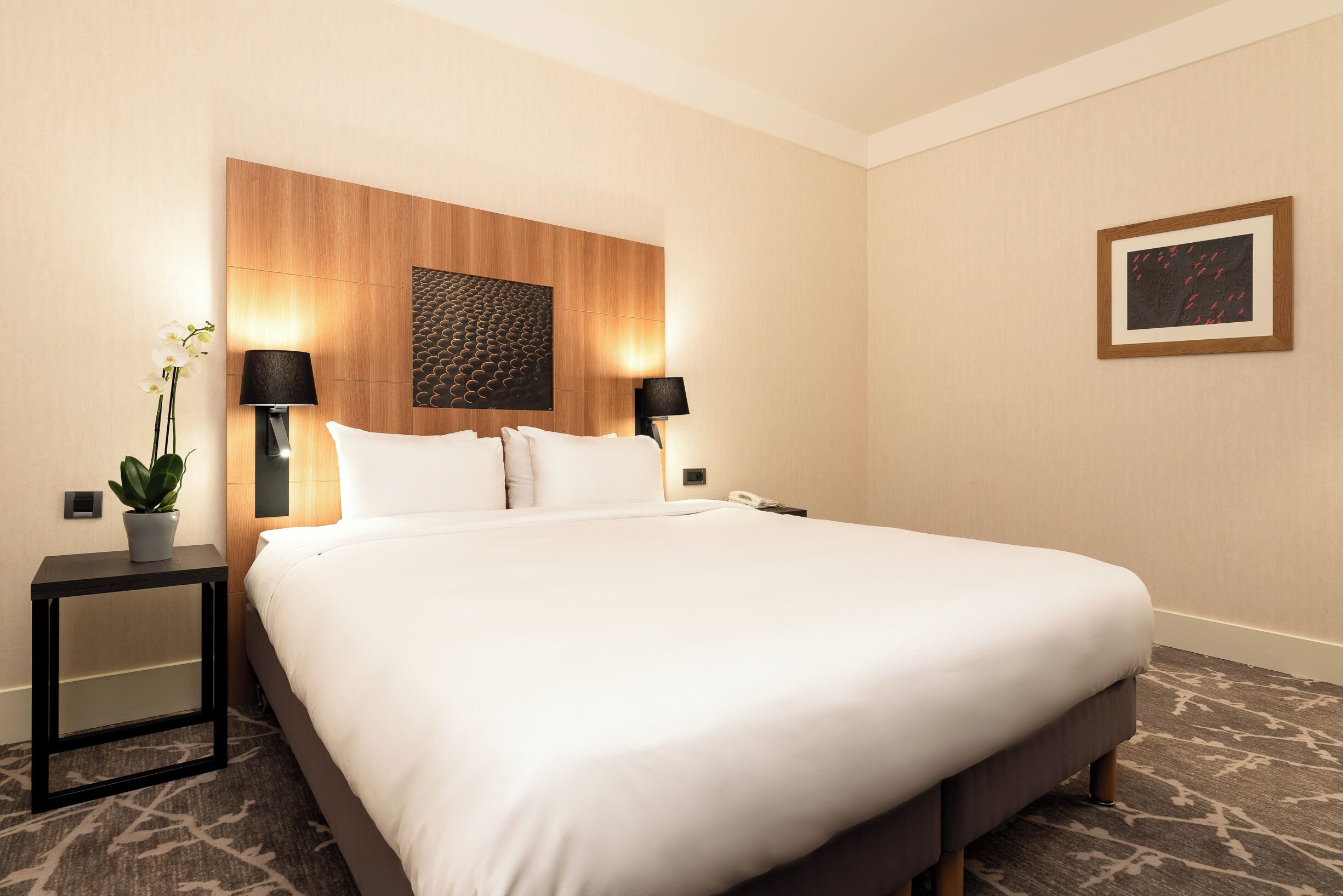 A hotel room with a large bed, two bedside tables, a potted plant, and a framed picture on the wall.