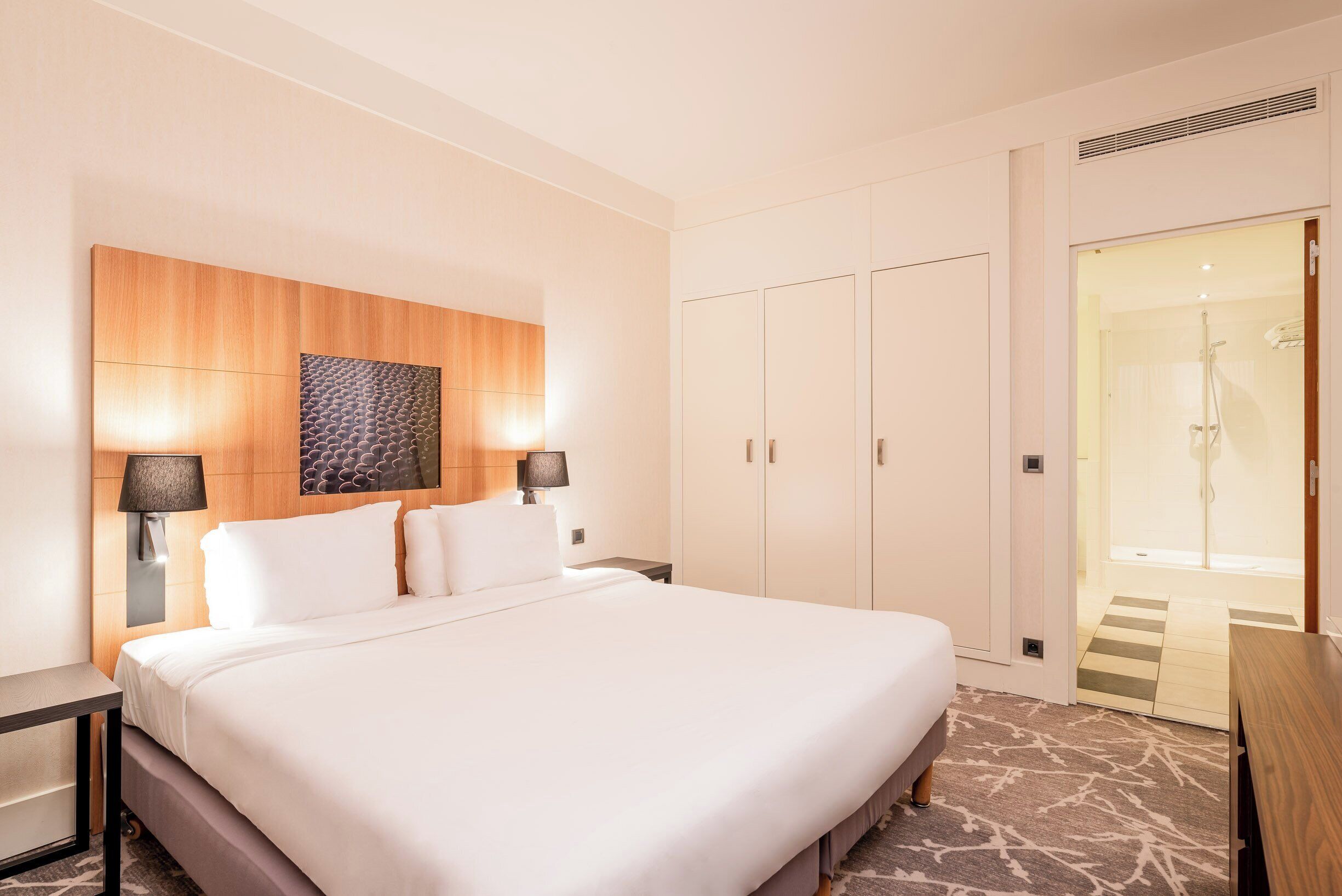 Ett hotellrum med en stor säng, en sänggavel i trä, två väggmonterade lampor, en garderob och en duschhörna.