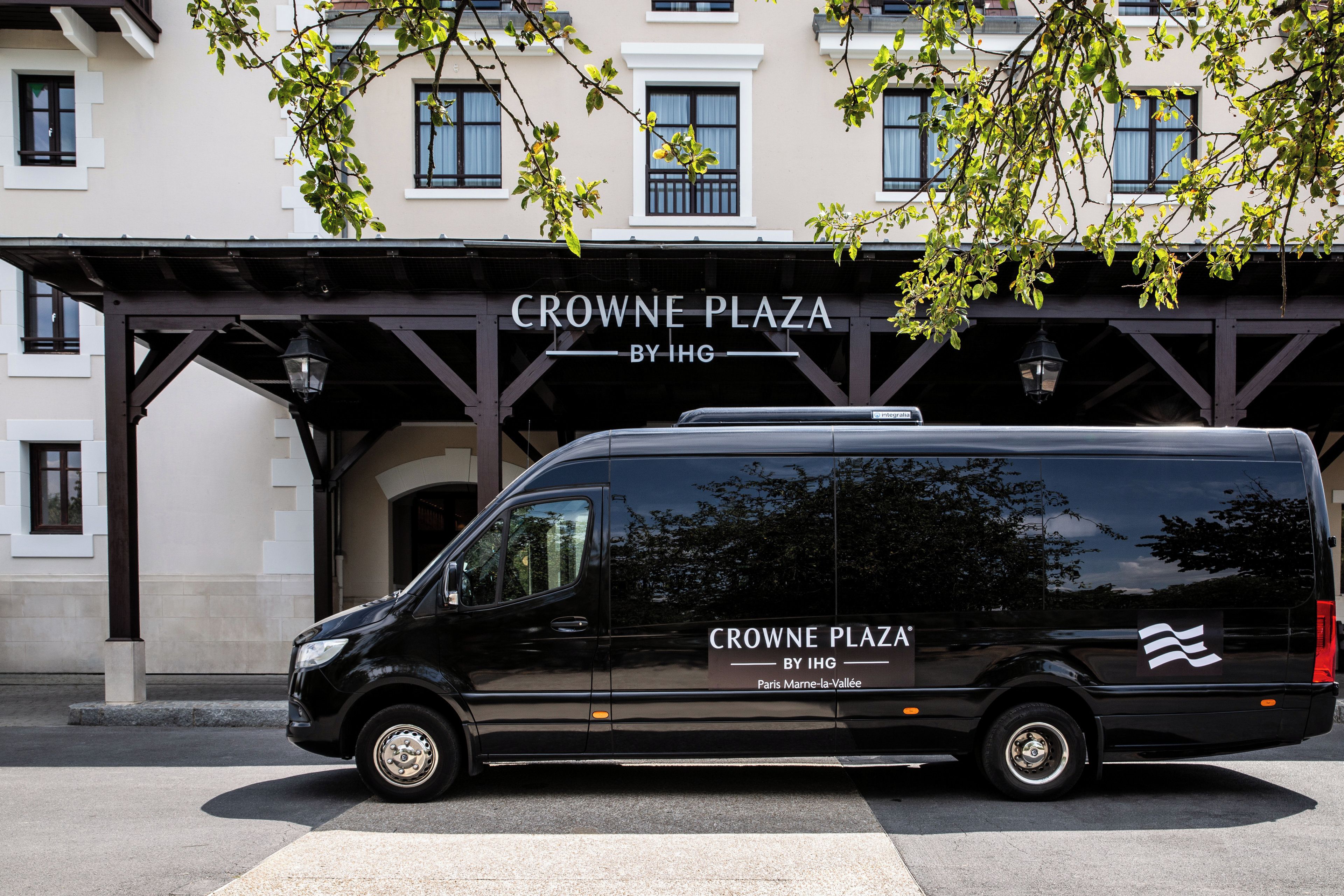 Ein schwarzer Van parkt vor einem Gebäude, an dem ein Schild mit der Aufschrift 'CROWNE PLAZA BY IHG Paris Marne-la-Vallée' angebracht ist.