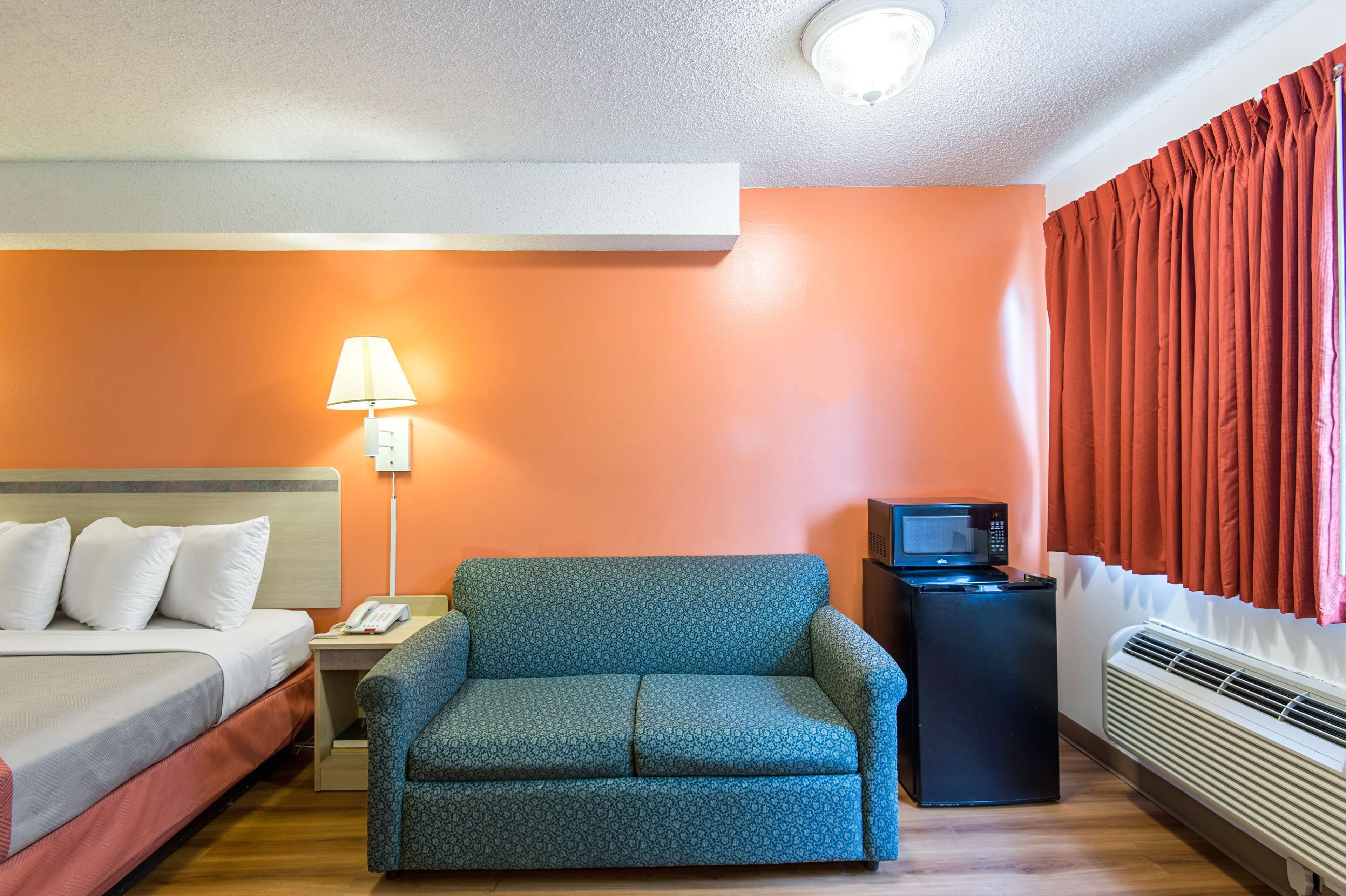 Quarto de hotel com uma cama, um sofá azul, uma escrivaninha com uma luminária, micro-ondas e uma janela com cortinas.