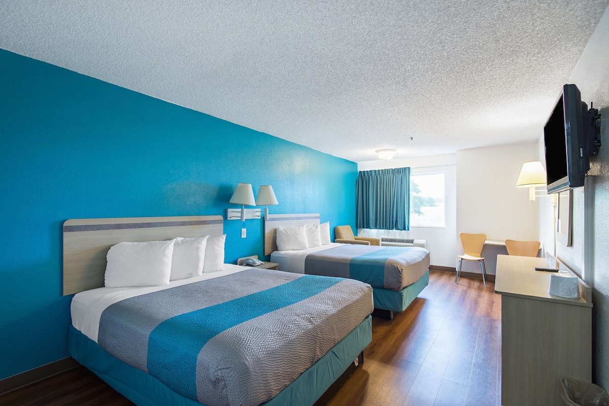 Quarto de hotel com duas camas, uma escrivaninha, uma cadeira, uma televisão e uma janela com cortinas.