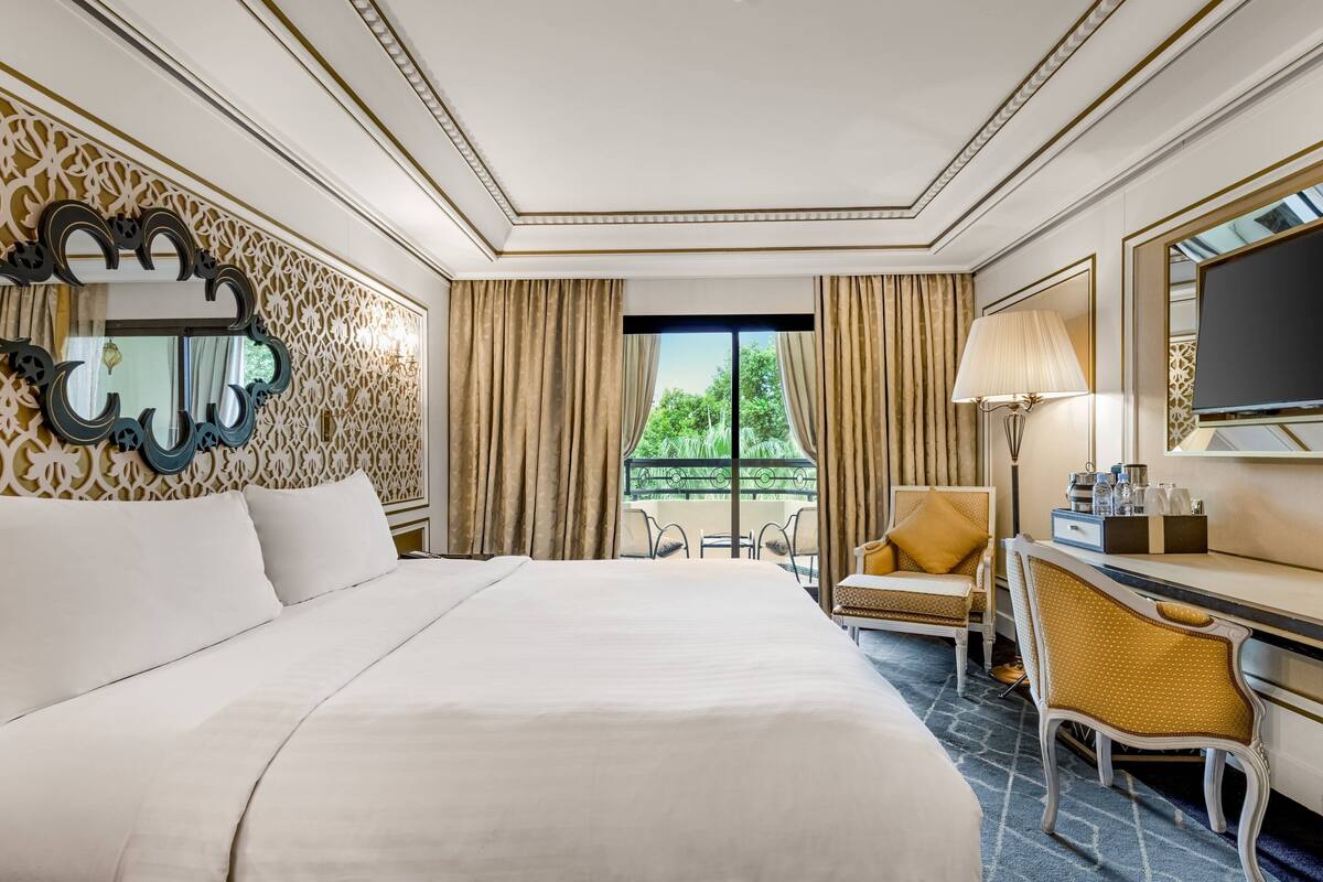 A hotel room with a large bed, a desk with a chair, a TV, and a view of greenery outside.
