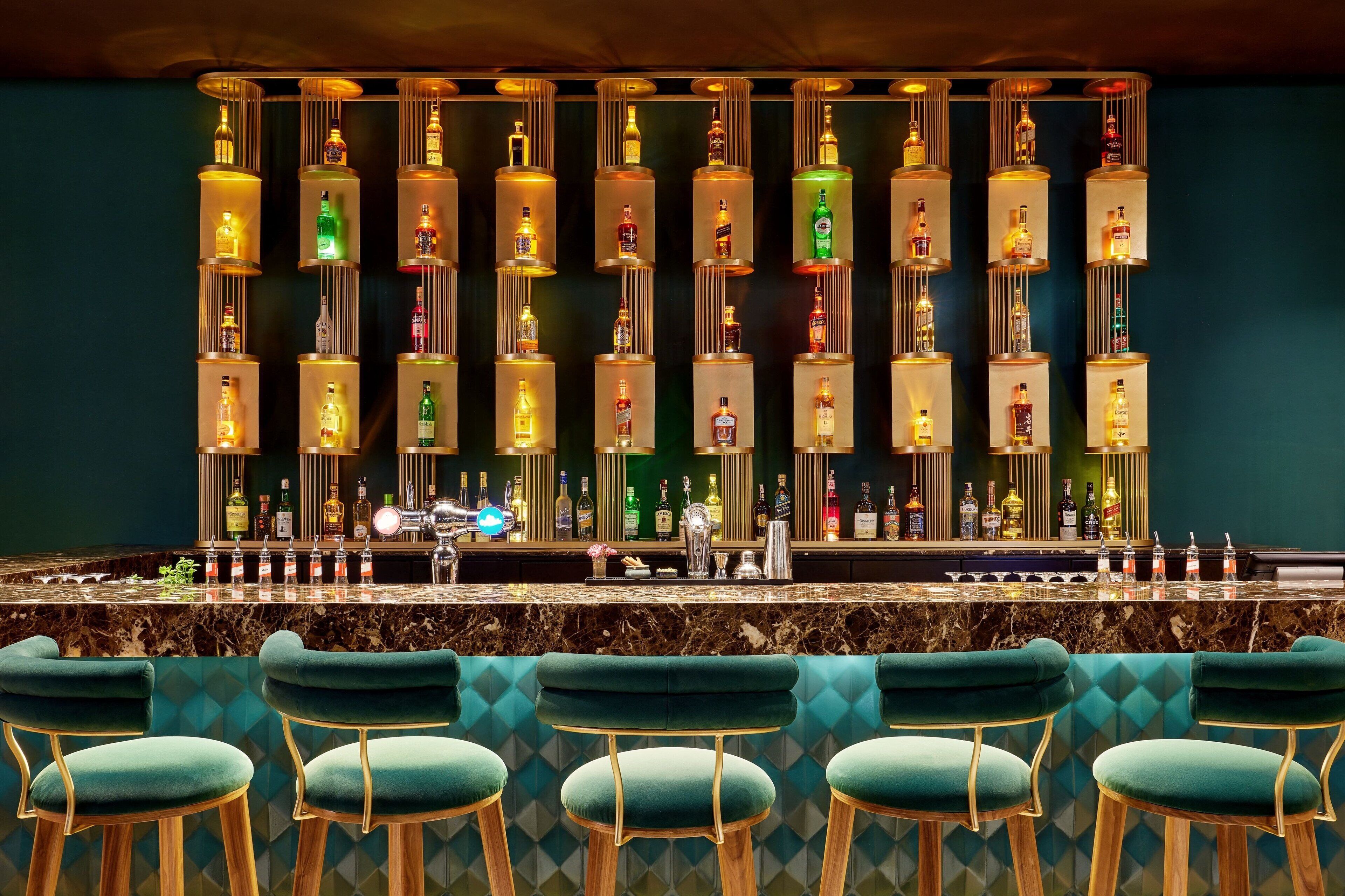 A bar area with a marble countertop, green upholstered bar stools, and a wall of illuminated liquor bottles.