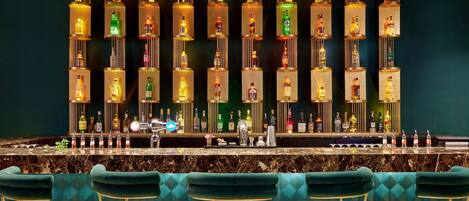 A bar area with a marble countertop, green upholstered bar stools, and a wall of illuminated liquor bottles.