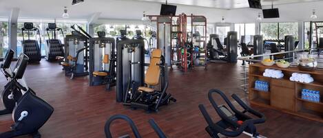 Un gimnasio amplio con diversa maquinaria de cardio y entrenamiento de fuerza, ventanas grandes y suelo de madera.