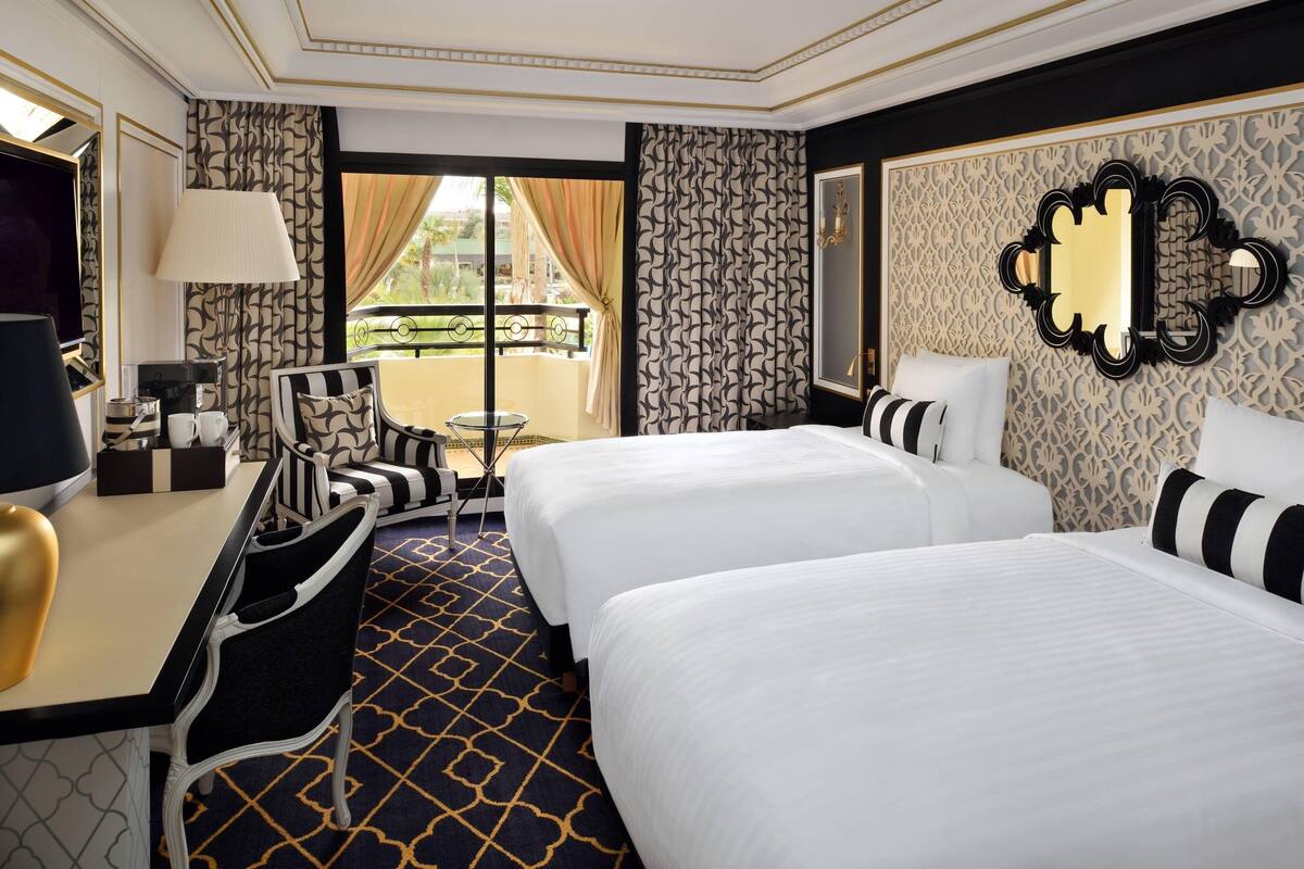 A hotel room with two beds, a desk with a chair, a TV, and a balcony with curtains.