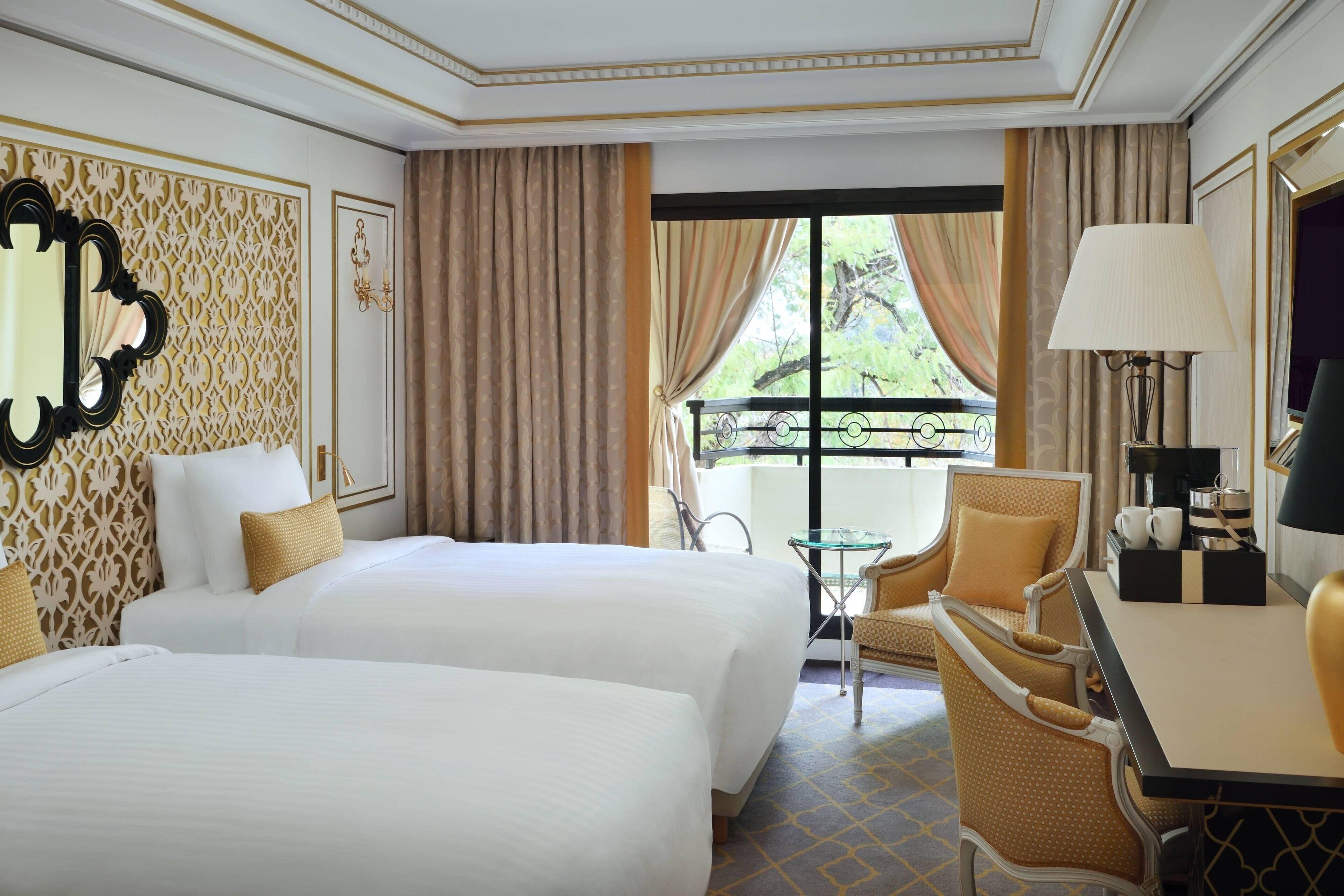 A hotel room with two beds, a small table, a chair, a lamp, and a balcony with curtains.