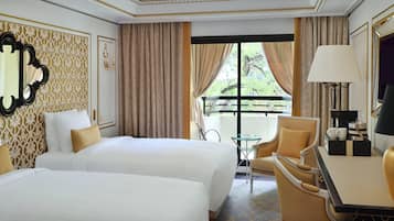A hotel room with two beds, a small table, a chair, a lamp, and a balcony with curtains.