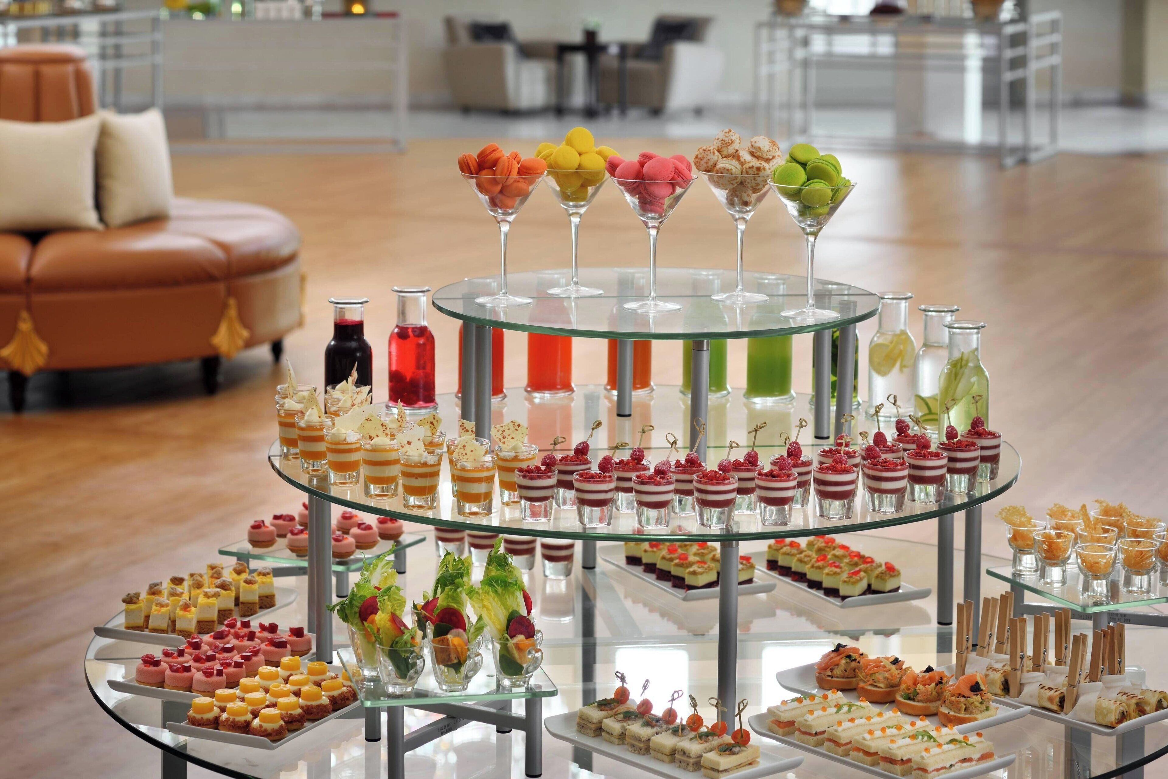 Eine mehrstufige Glasvitrine, gefüllt mit verschiedenen Desserts und Getränken, in einem modernen Ambiente mit einer Ledersofa und einem Essbereich im Hintergrund.