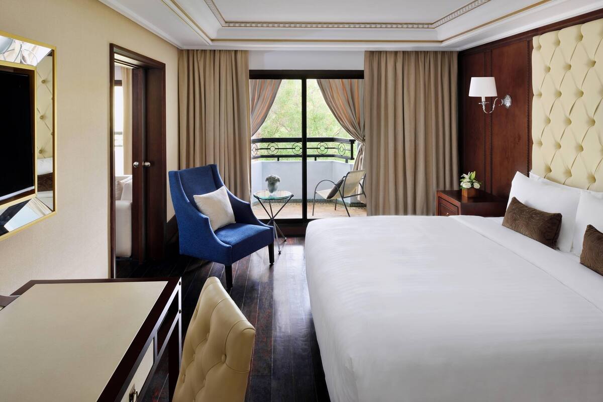 A hotel room with a large bed, a blue armchair, a small table, and a balcony with a view.