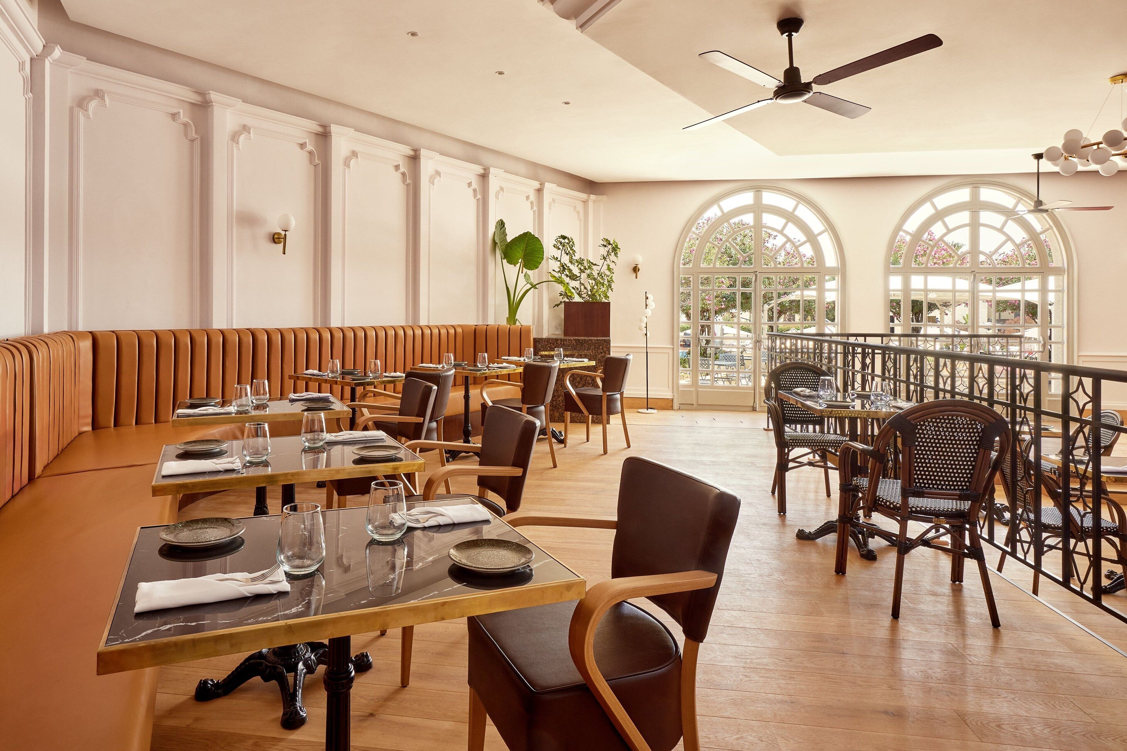 Un vaste espace dédié à la salle à manger, avec de grandes tables en bois et des banquettes rembourrées, de grandes fenêtres surmontées d’arcs, et un balcon doté d’une balustrade.