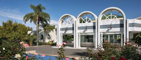 A hotel entrance with arched windows, a pool, and surrounding greenery.