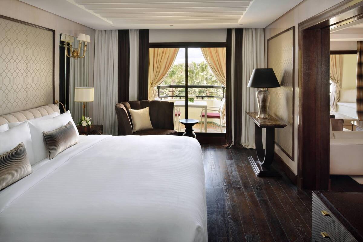 A hotel room with a large bed, a sitting area with a table and chairs, a balcony with curtains, and a view of palm trees.
