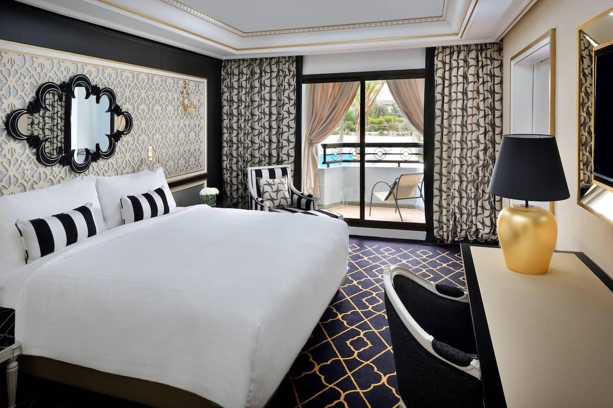 A hotel room with a large bed, a desk with a lamp, a chair, a balcony with a view, and patterned wallpaper.