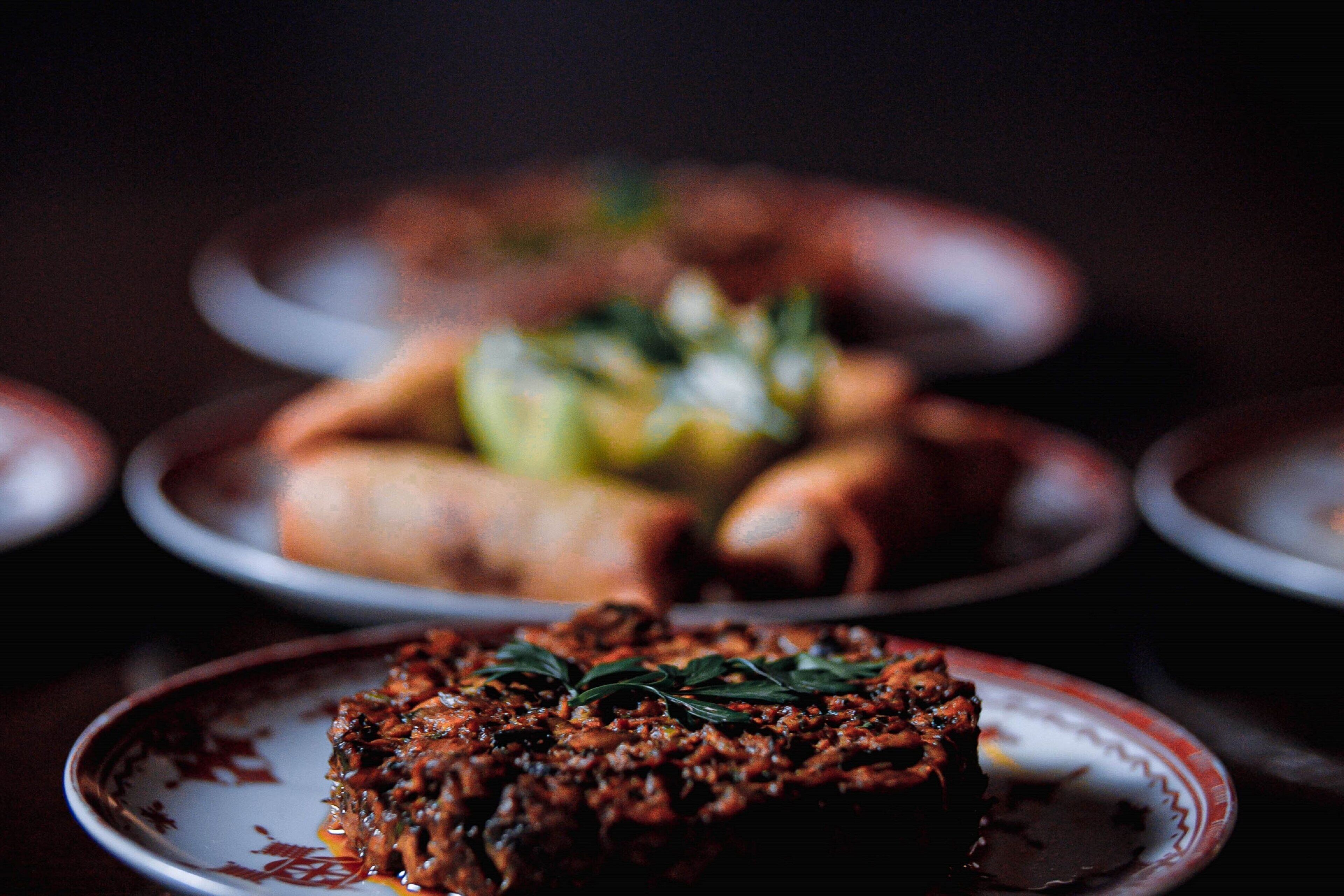 A close-up of a dish with a garnish on a plate, with blurred dishes in the background.