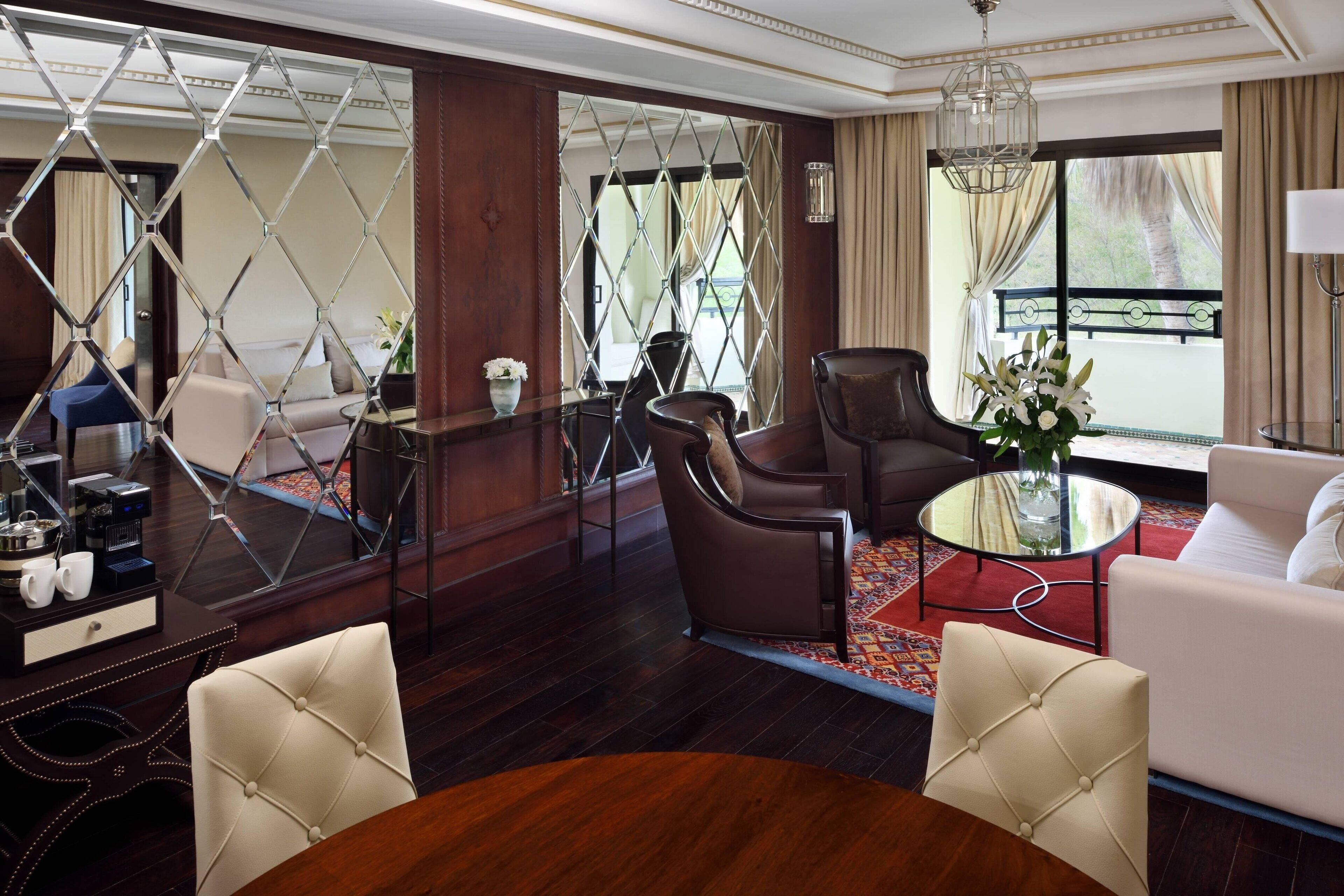 A hotel room with a wooden floor, a round dining table, two armchairs, a sofa, a coffee table, a decorative mirror, and a view of the outdoors through large windows.