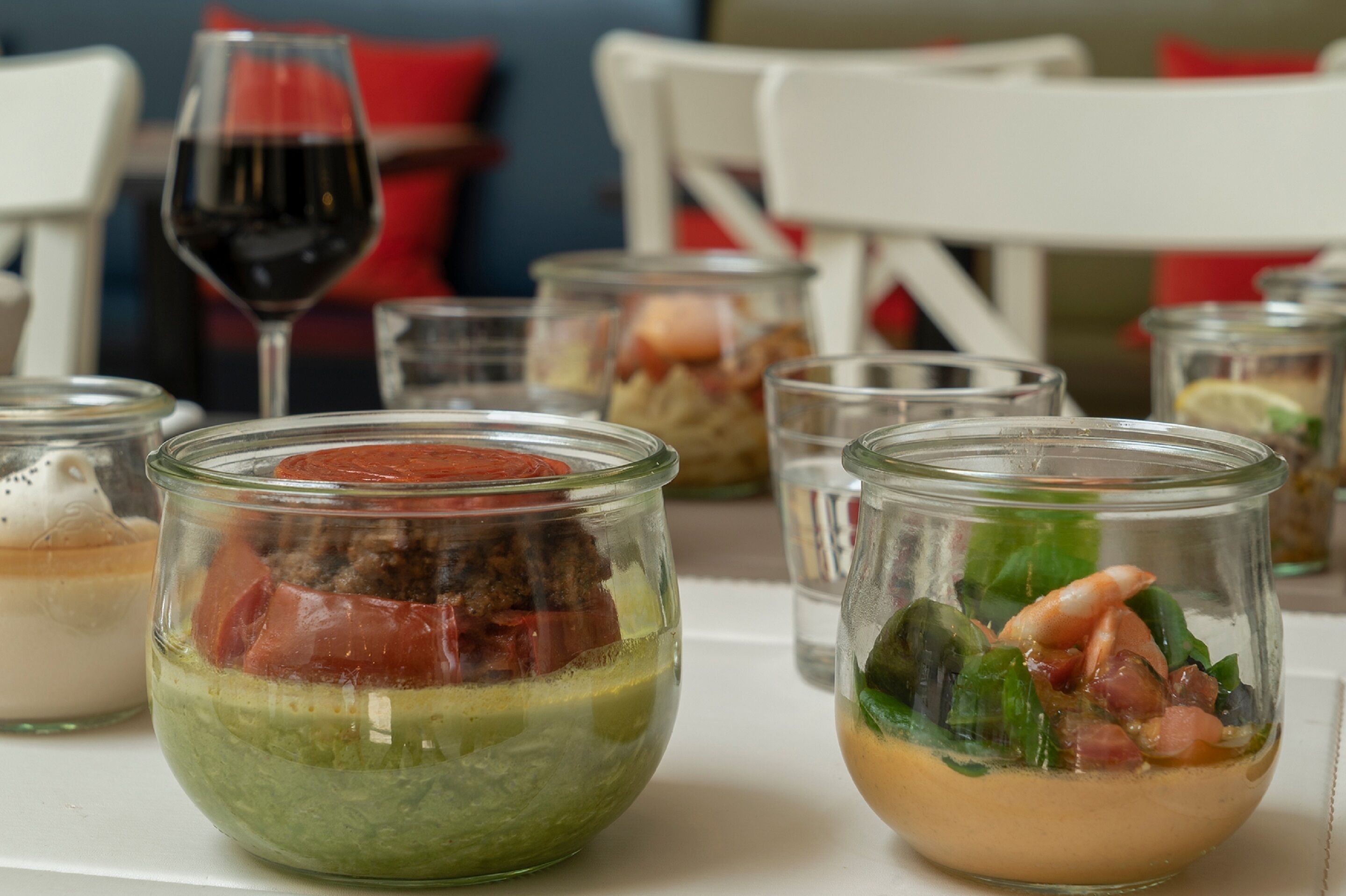 Une table à manger avec divers récipients en verre contenant des aliments, un verre à vin et une chaise blanche en arrière-plan.