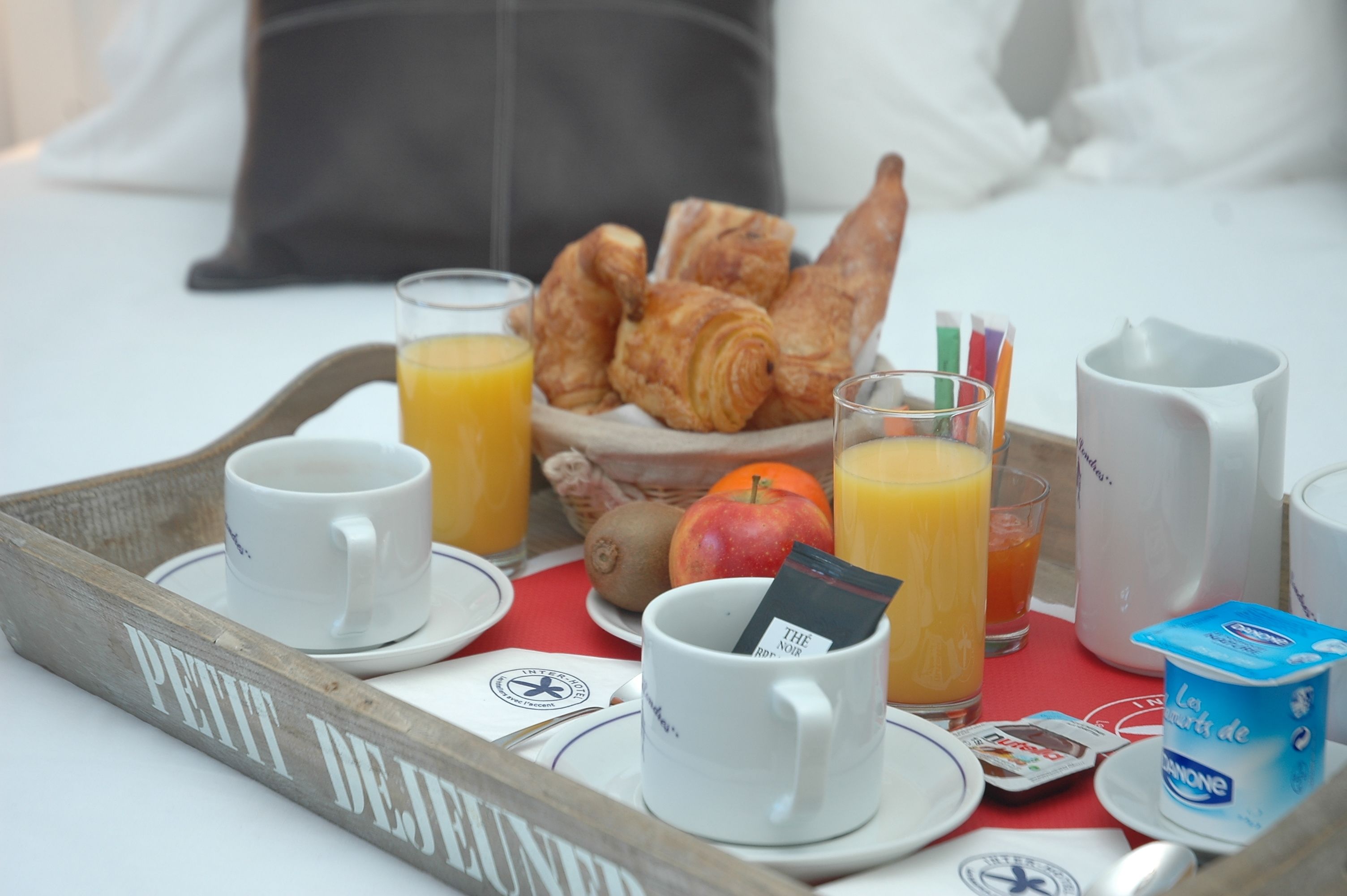 A breakfast tray with croissants, juice, a mug, and a small container of yogurt on a bed with pillows.